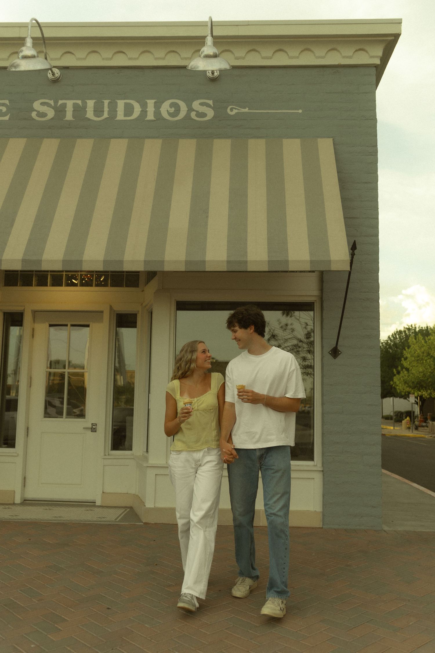 A young couple walks hand in hand outside a studios storefront with striped awning on a brick sidewalk.
