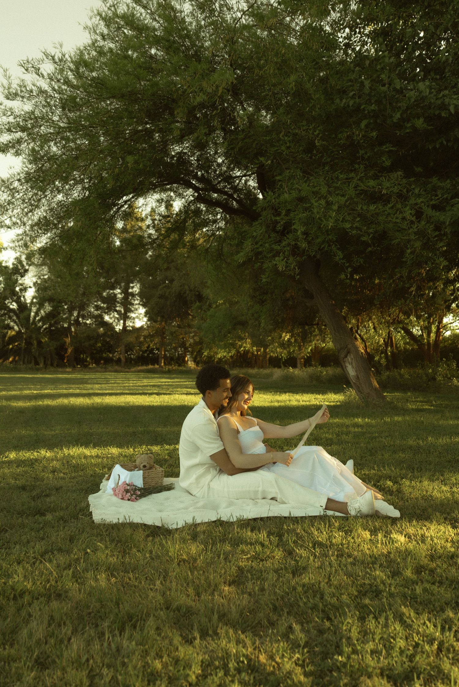 Couple in white sitting on blanket under tree during golden hour maternity photoshoot in park.