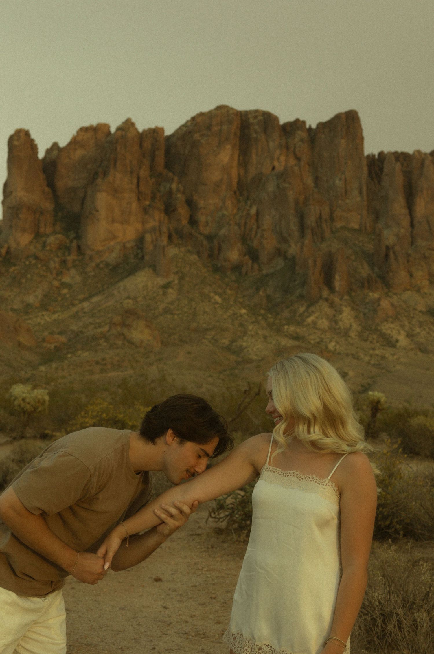 A man kissing a woman's hand in a romantic desert setting with dramatic red rock formations in the background.