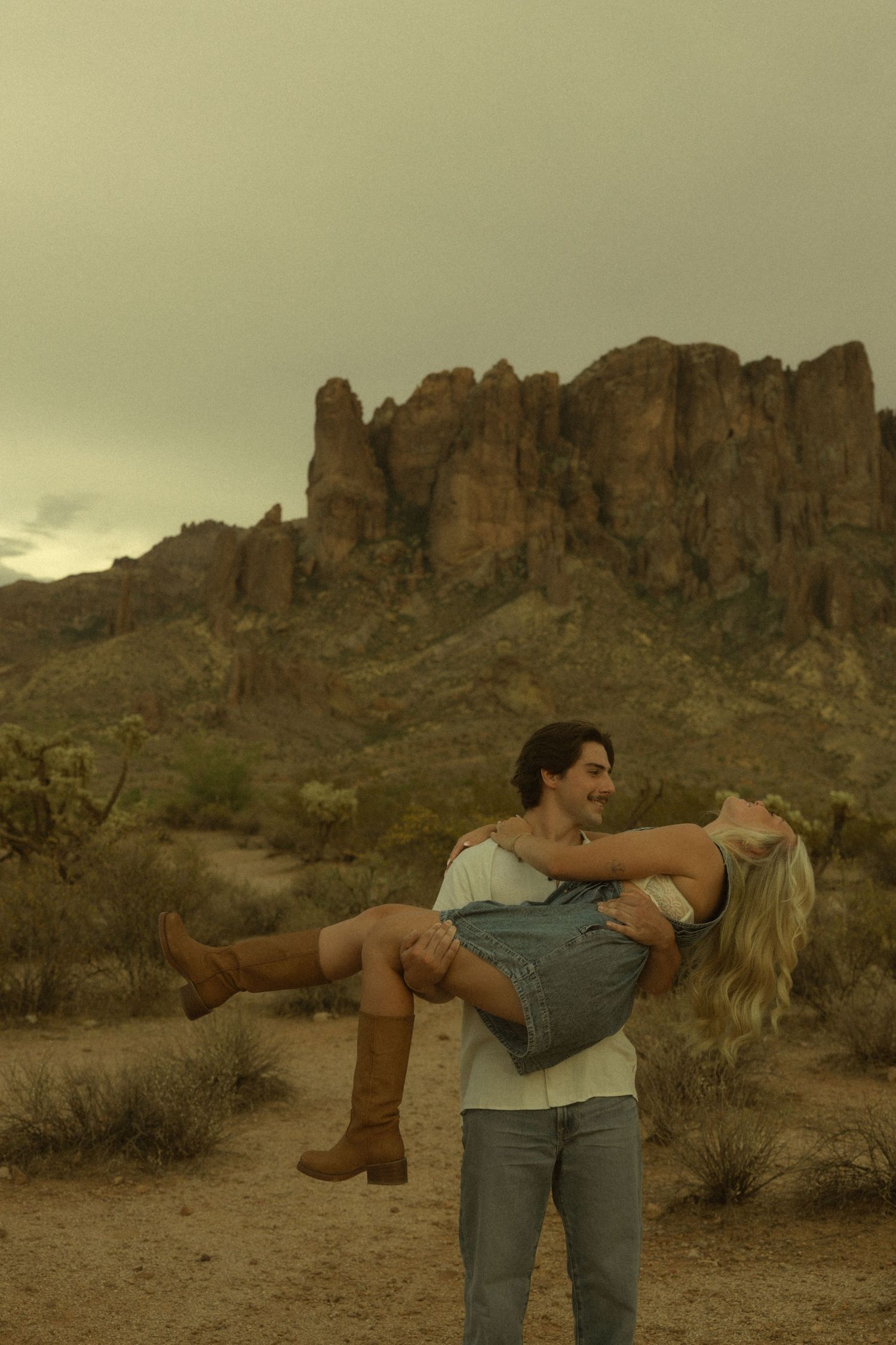 A couple poses romantically in the desert, man carrying woman in his arms with rugged rock formations behind them.