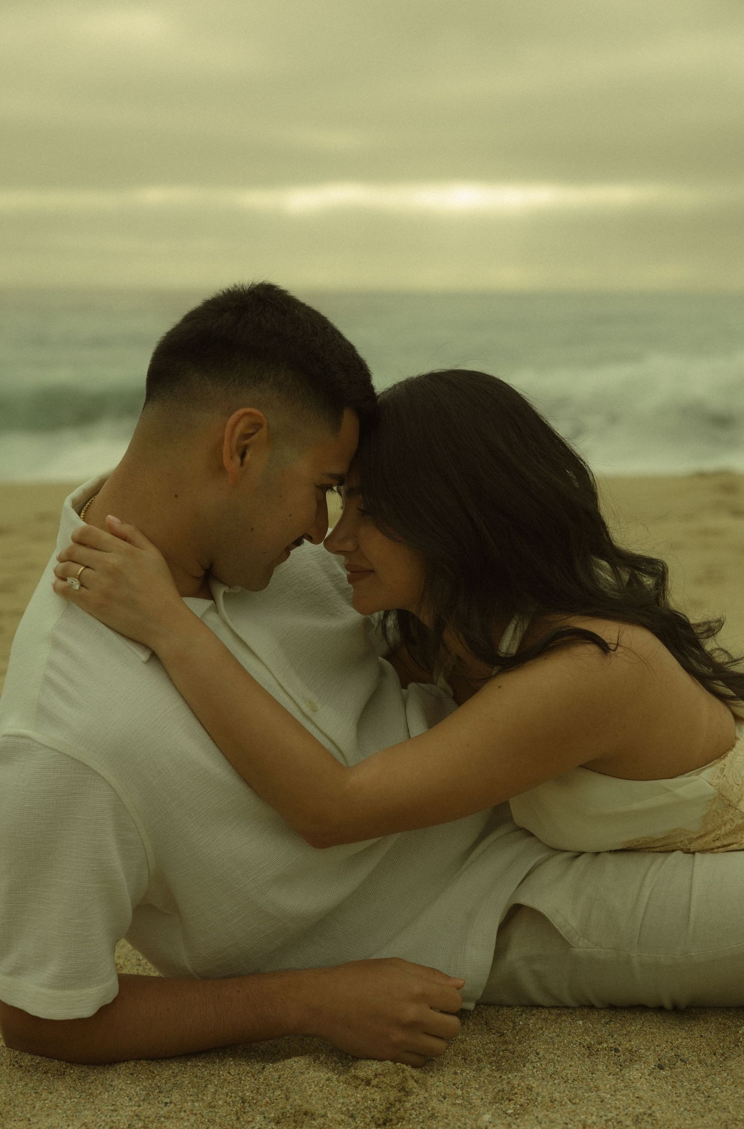 A romantic couple sits close together on a sandy beach at sunset, foreheads touching tenderly.