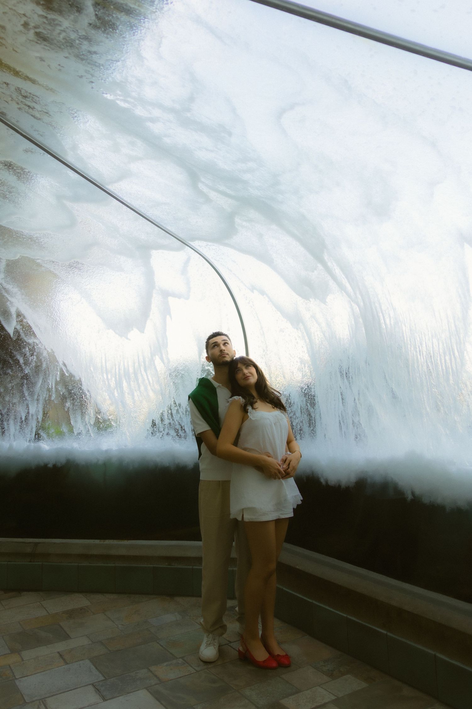 A couple embraces in front of a large indoor waterfall feature, woman in white dress and man in green shirt.