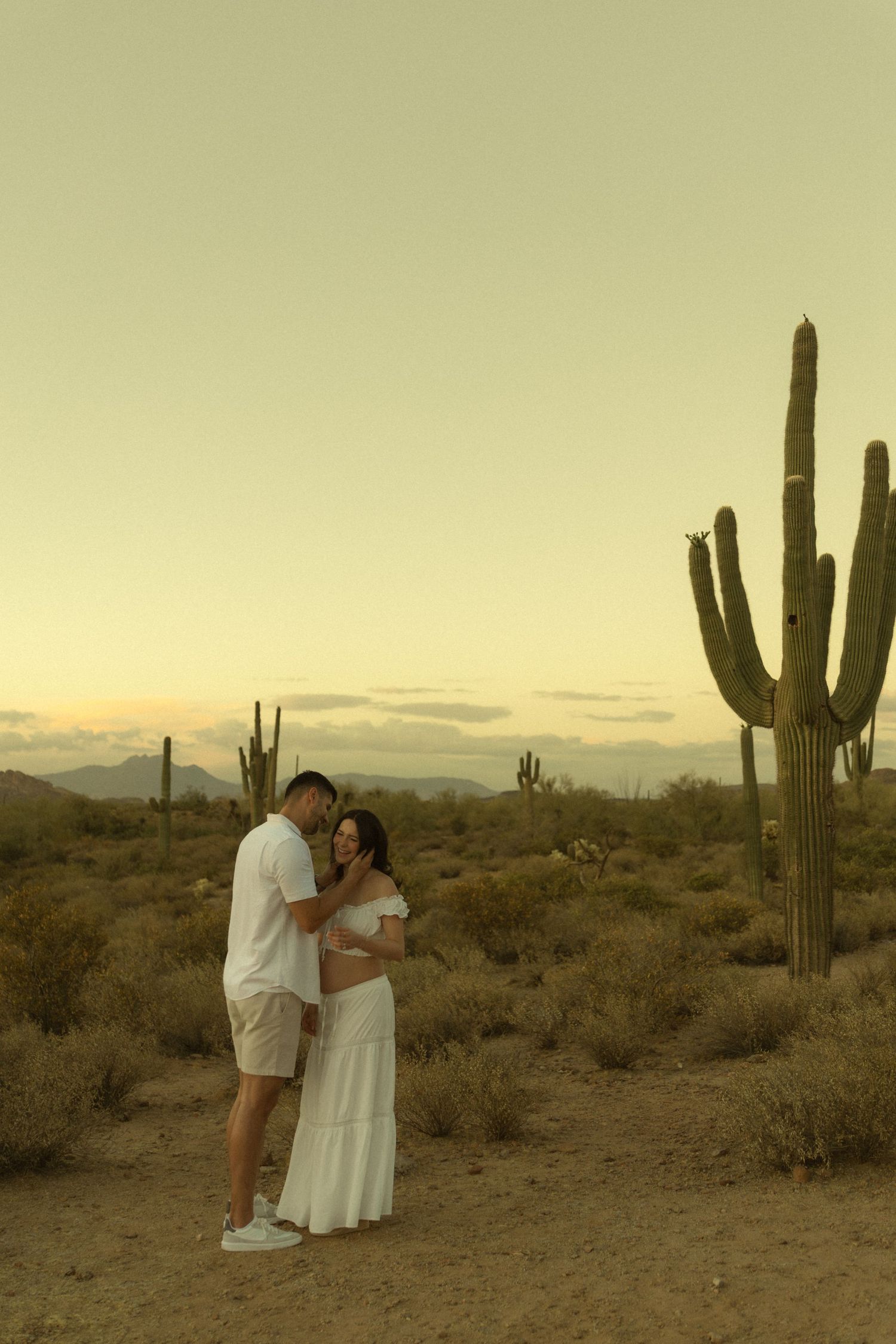Couple in white outfits embracing in Sonoran Desert at sunset with tall saguaro cacti in background.