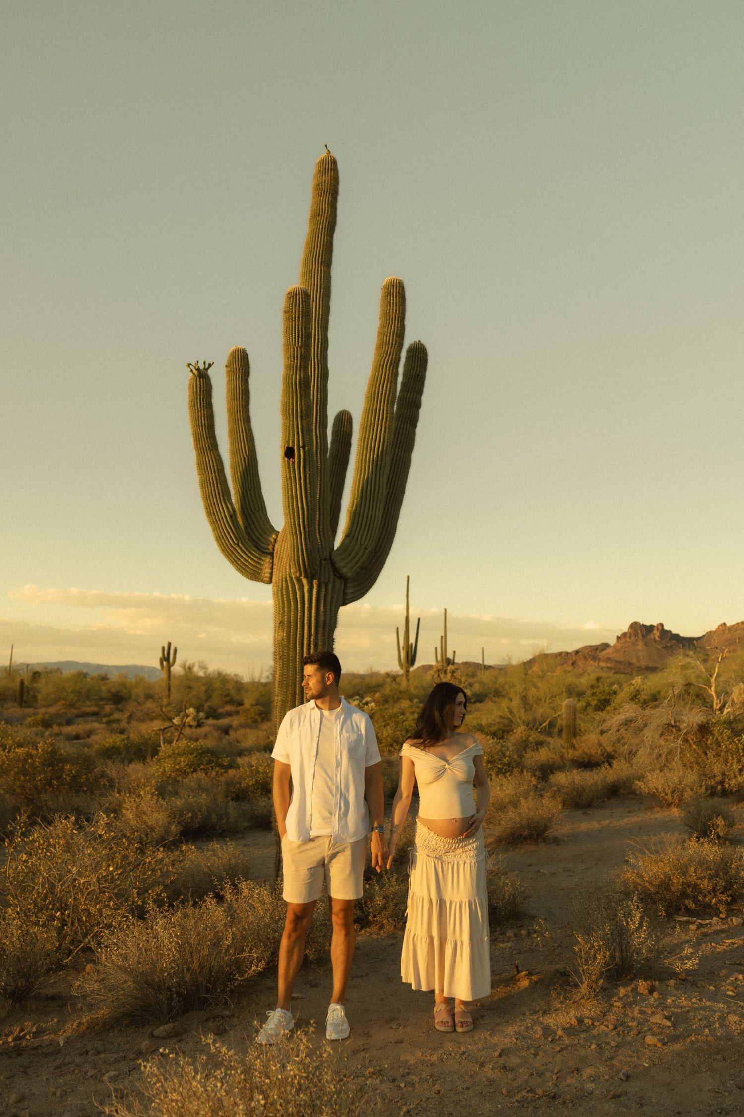 Couple holding hands in white outfits standing before a tall saguaro cactus in the Sonoran Desert at golden hour.