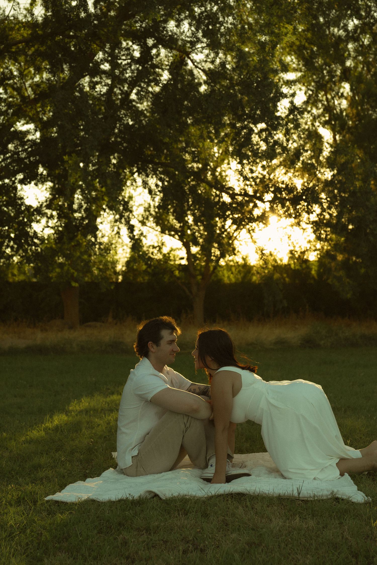 Couple in white sitting on blanket in lush green park at golden sunset, sharing romantic moment outdoors.