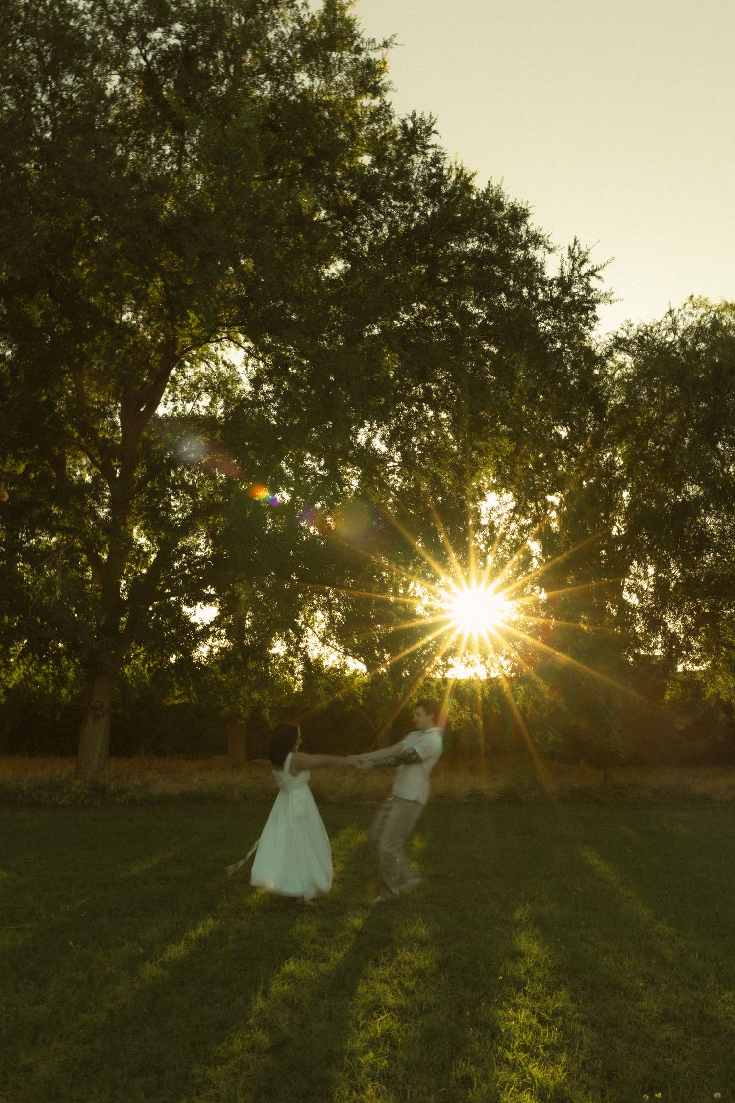 Couple dancing in a sunlit green field at sunset with trees and golden sun rays in the background.