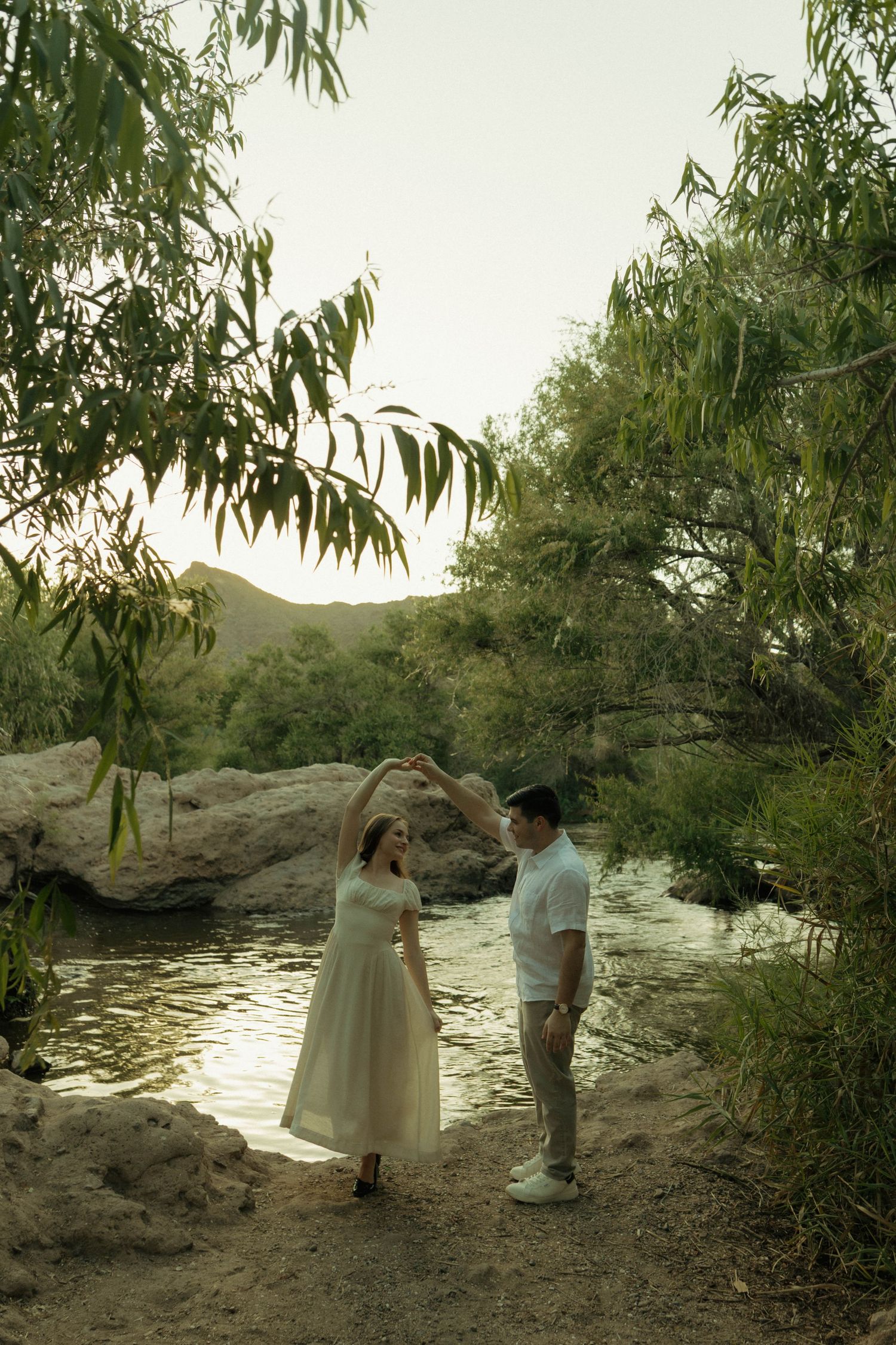 Couple dancing near a sunlit creek surrounded by lush green trees and rocks in a serene outdoor setting.