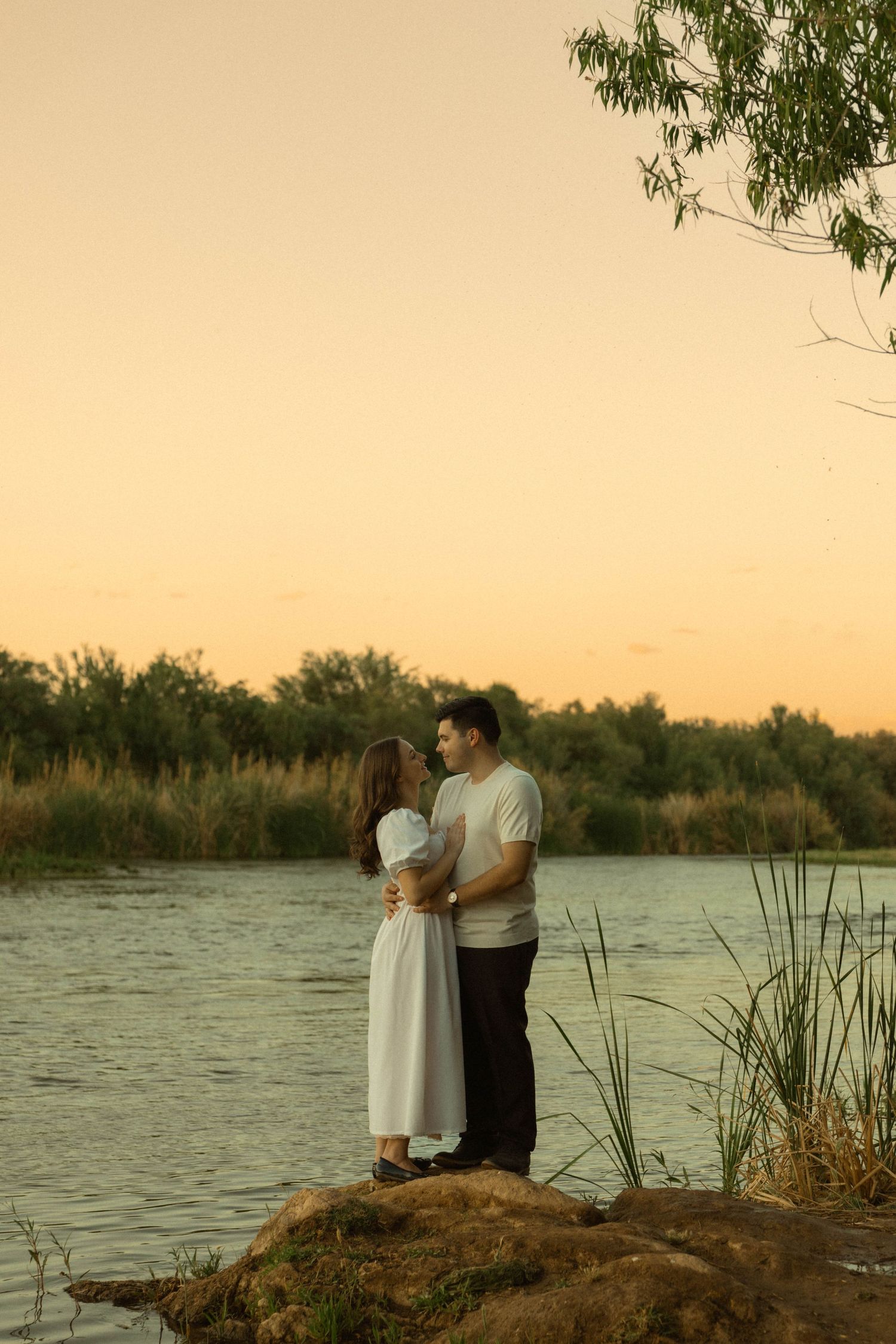 Romantic couple standing together on rocks by a calm river at golden sunset, woman in white dress.