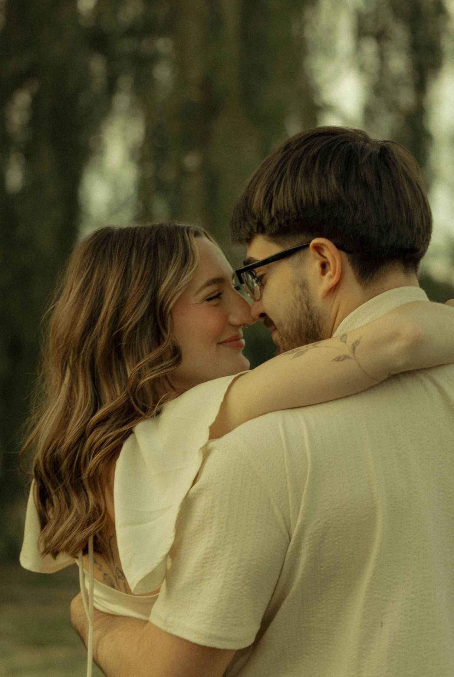 A smiling couple embraces intimately outdoors, the woman with wavy hair and man wearing glasses in cream attire.