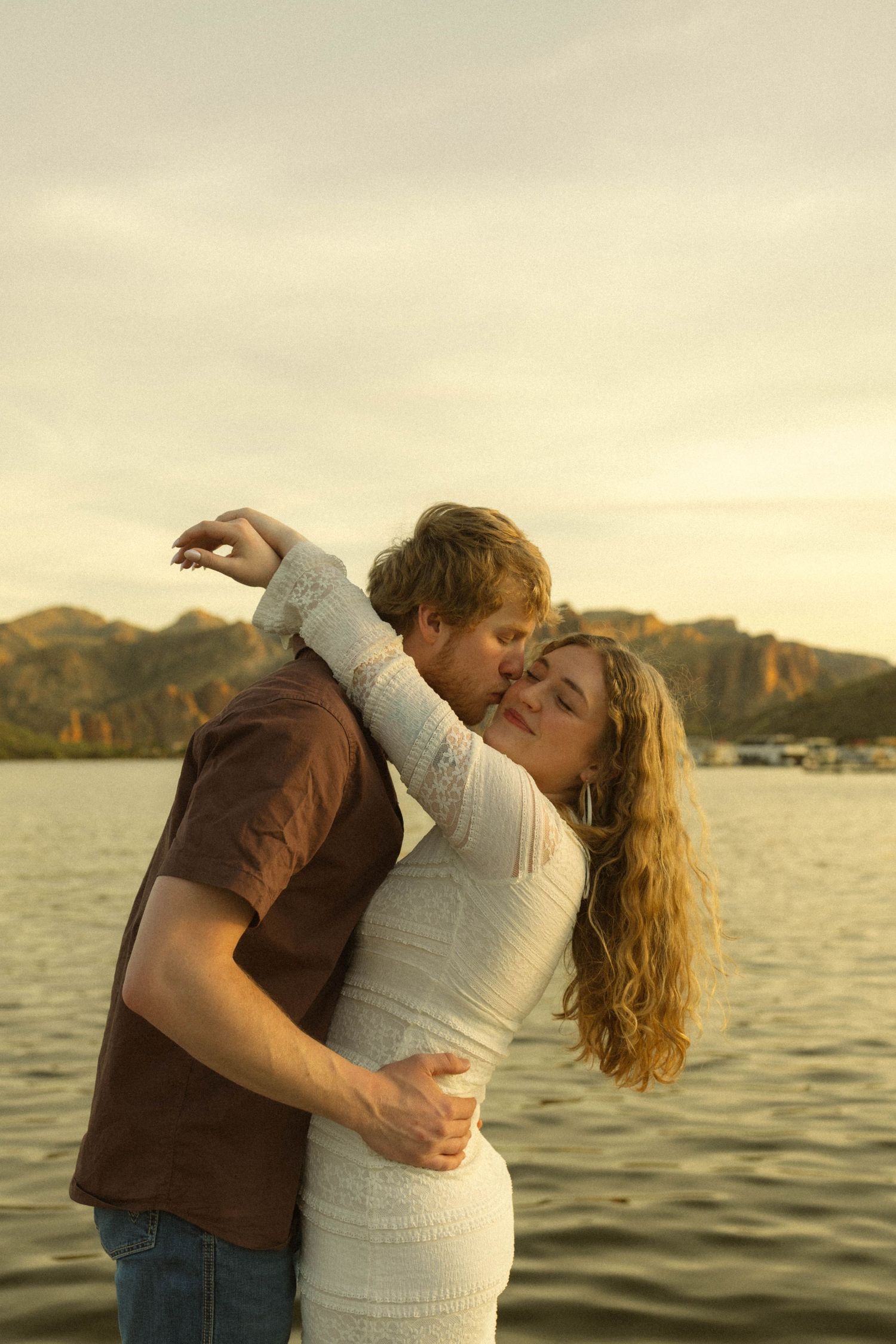 Couple embracing passionately by a lake at golden hour with mountains in the background.