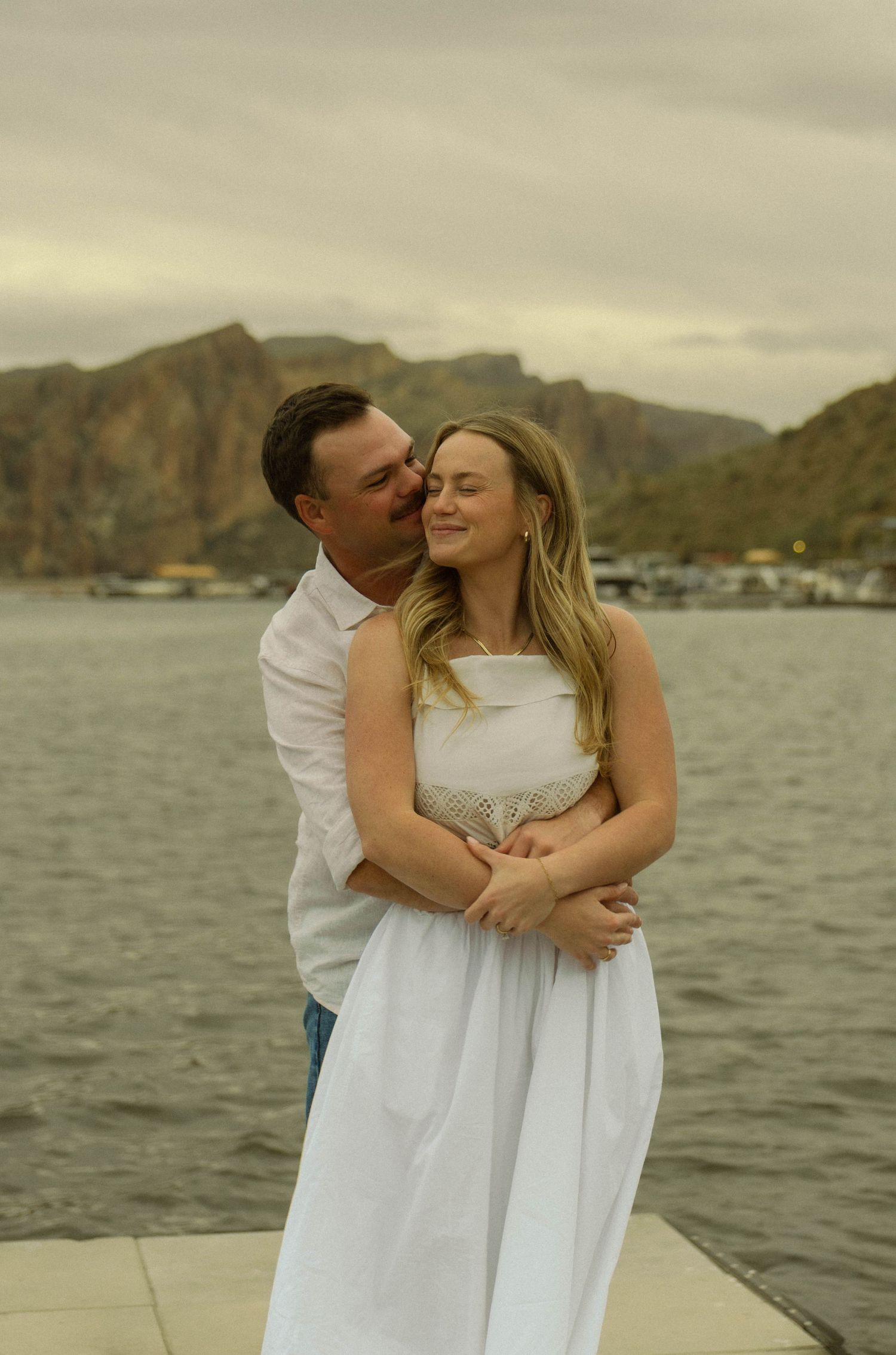 A couple in white clothing embraces on a dock with mountains and water in the background.