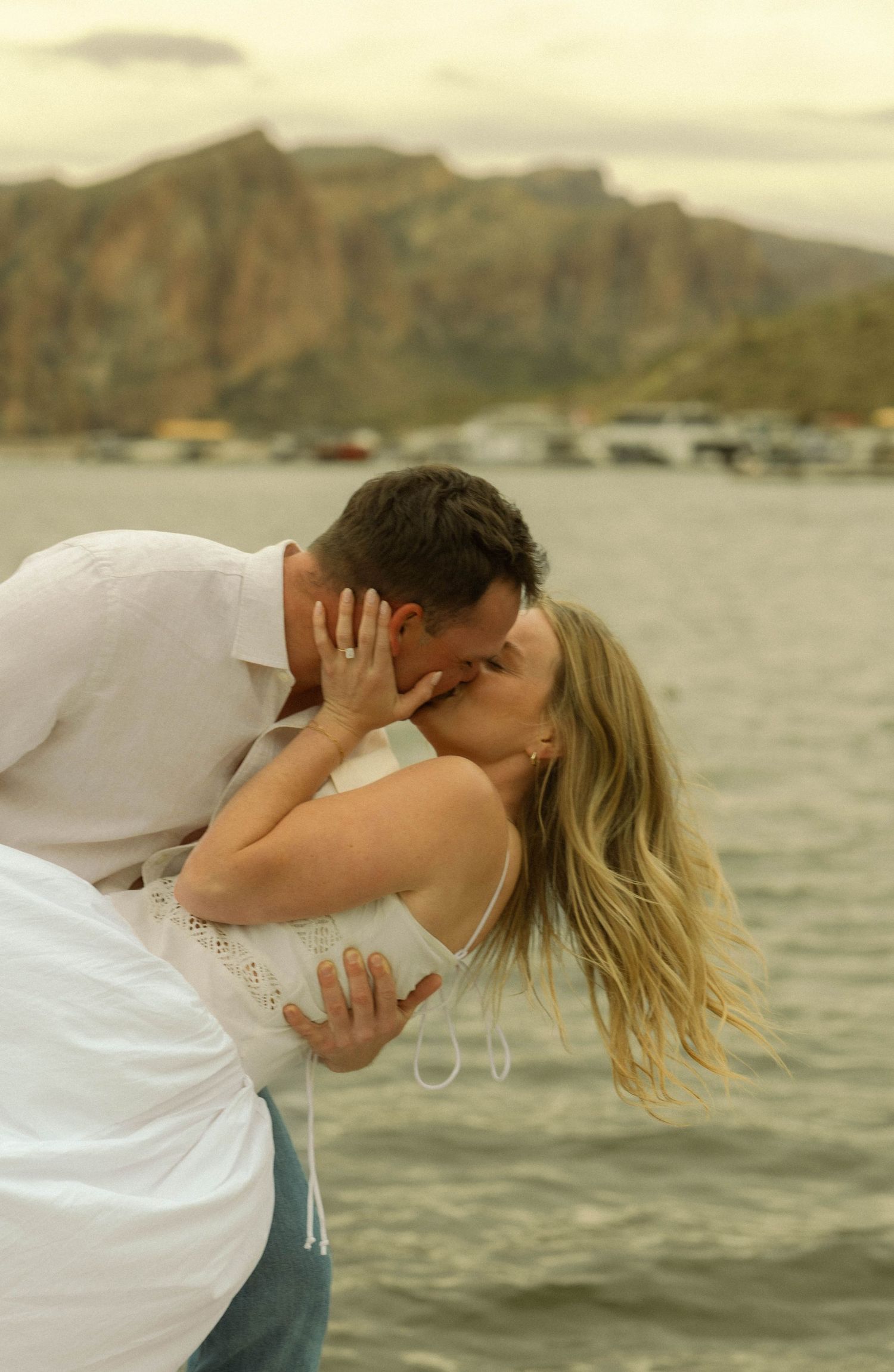 A loving couple shares a romantic kiss during a dip pose by the waterfront with mountains behind them.