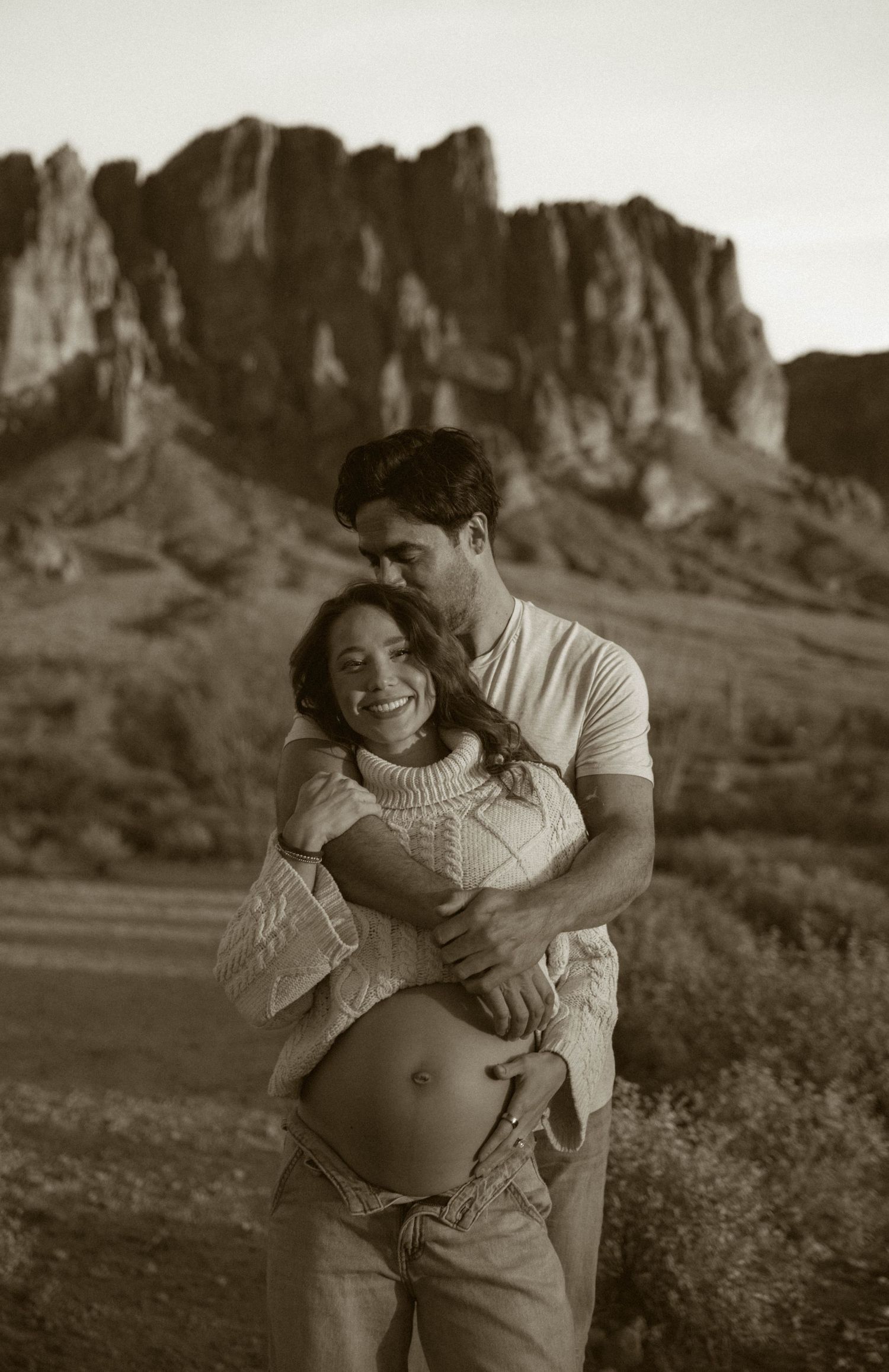 A couple embraces in a romantic maternity photo with dramatic mountain cliffs in the background at sunset.