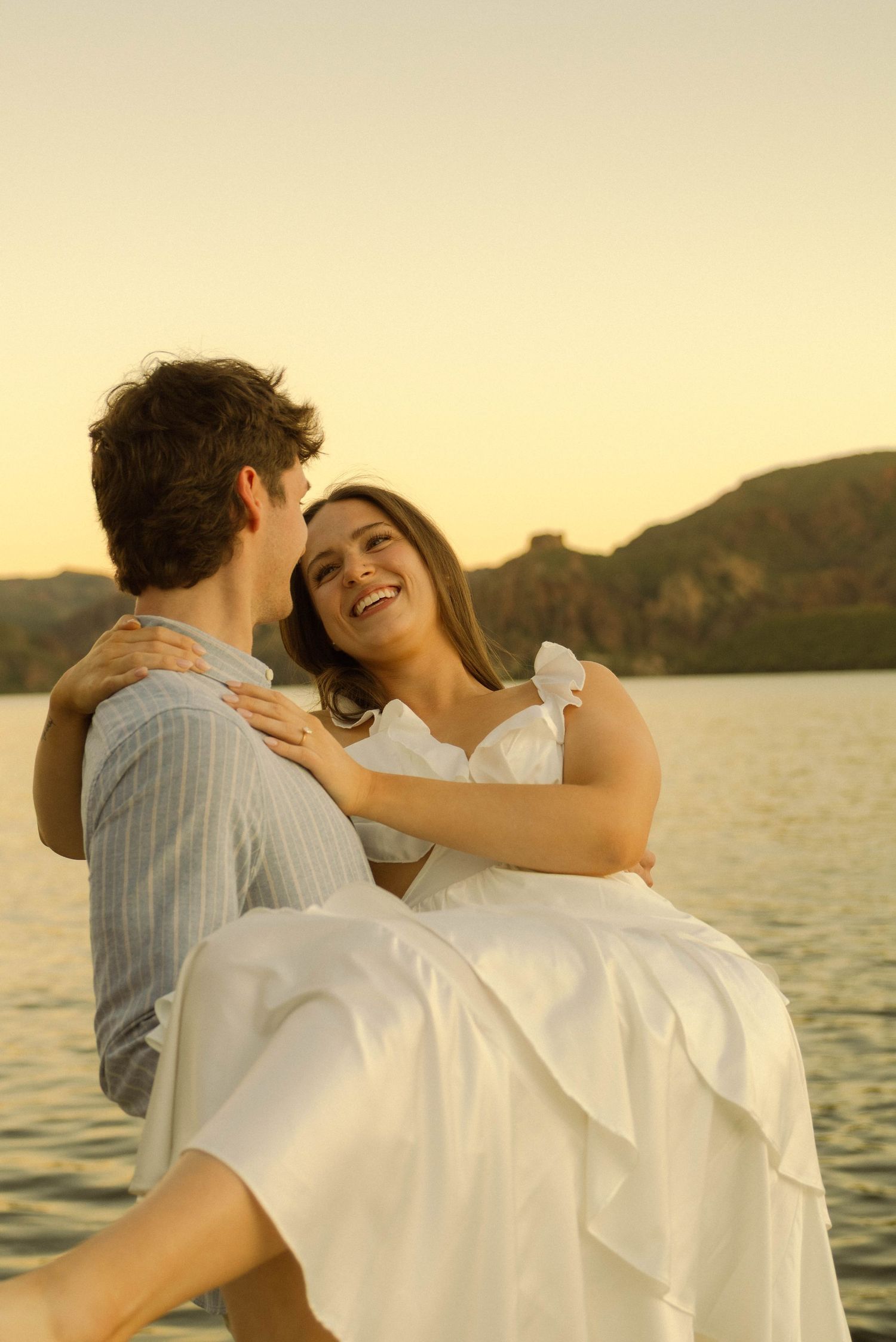 A romantic couple embraces during golden hour by a lake with mountains in the background.