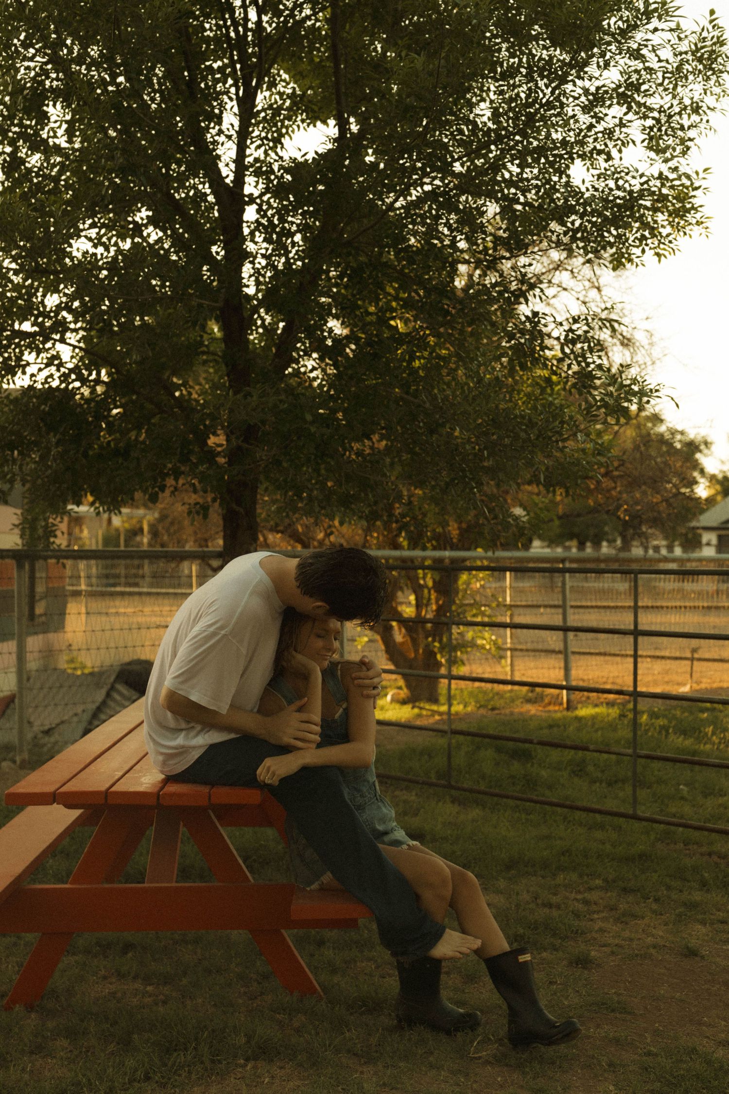 A couple shares a tender moment sitting together at a red picnic table under a tree at sunset.