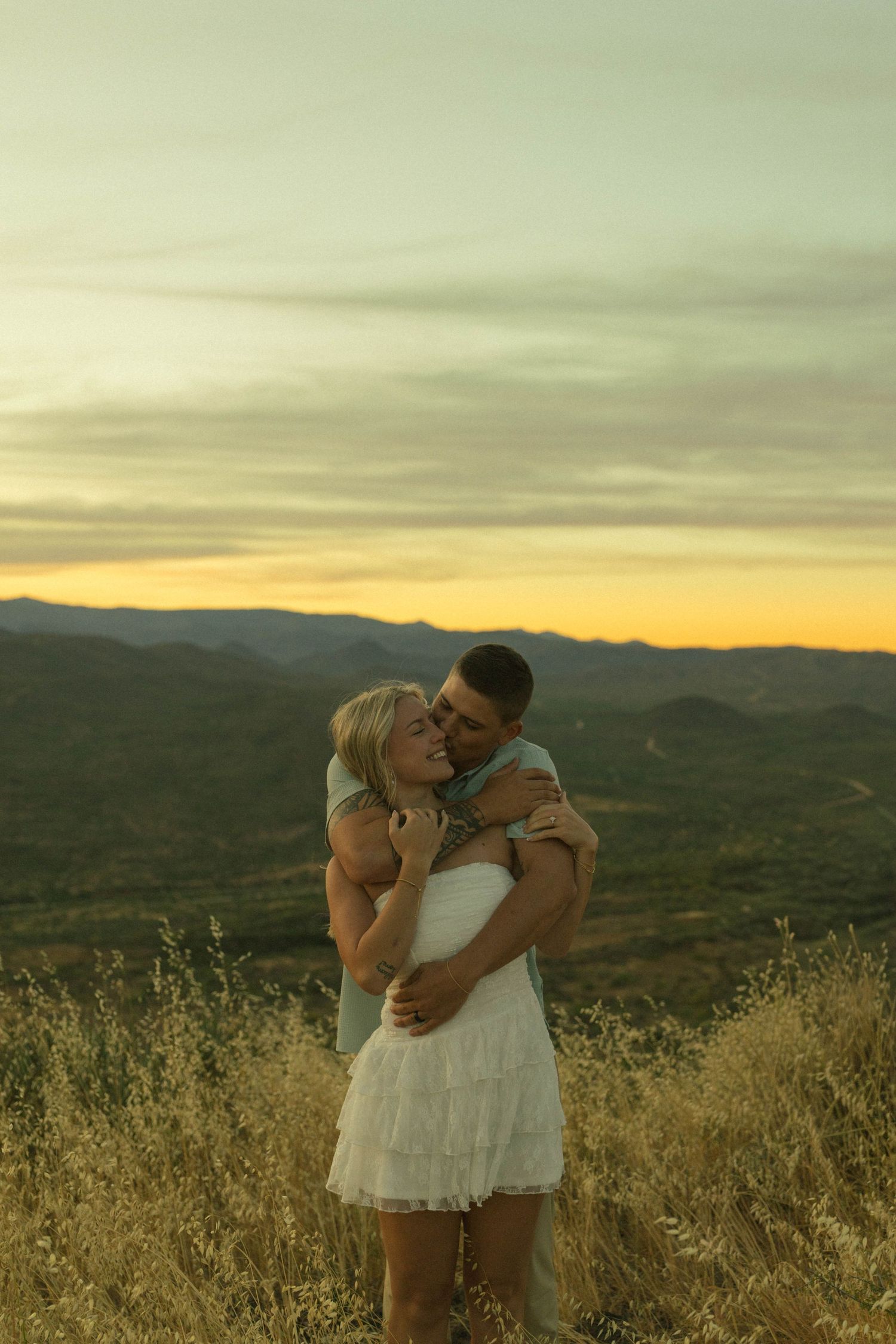 Romantic couple kissing at sunset in golden meadow with scenic mountain backdrop.