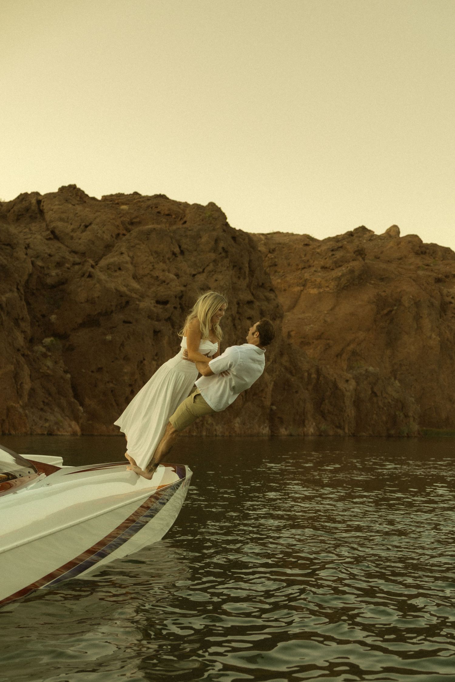 Man lifting woman in white dress on the bow of a speedboat with rugged rocky cliffs at golden hour.