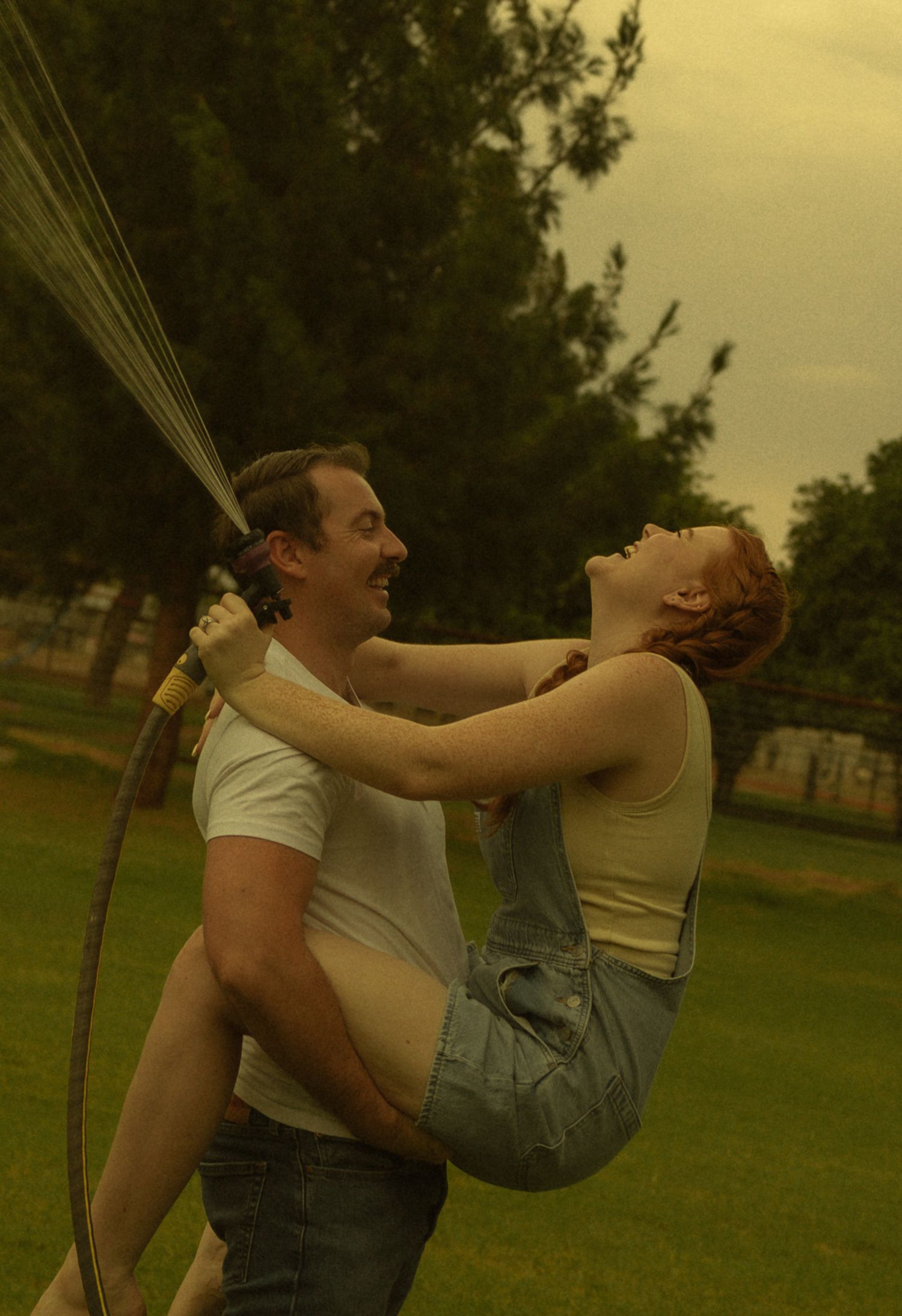 A man playfully holds a woman while spraying water from a garden hose outdoors on a warm day.