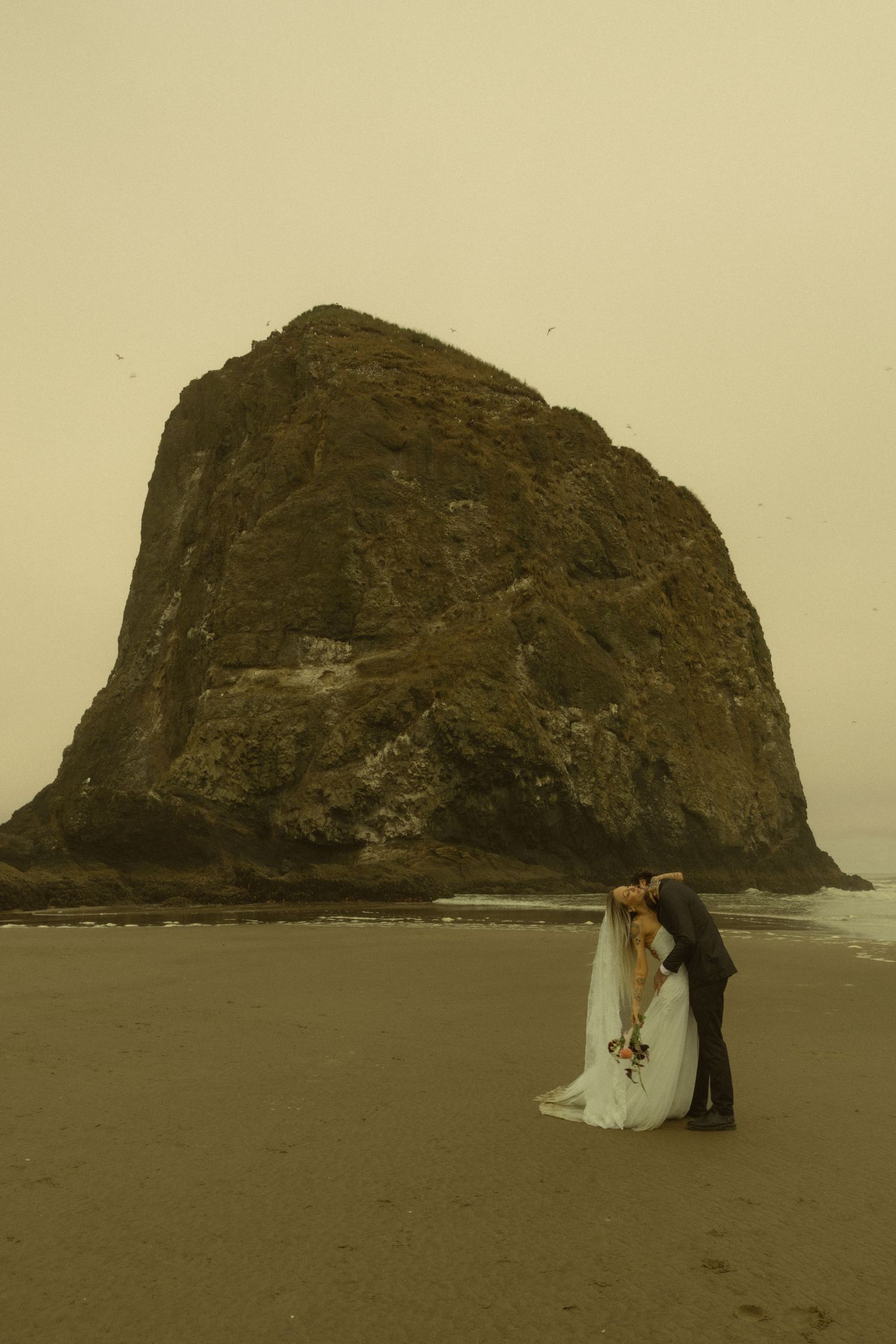 Bride and groom kissing on sandy beach with iconic Haystack Rock at Cannon Beach, Oregon in background.