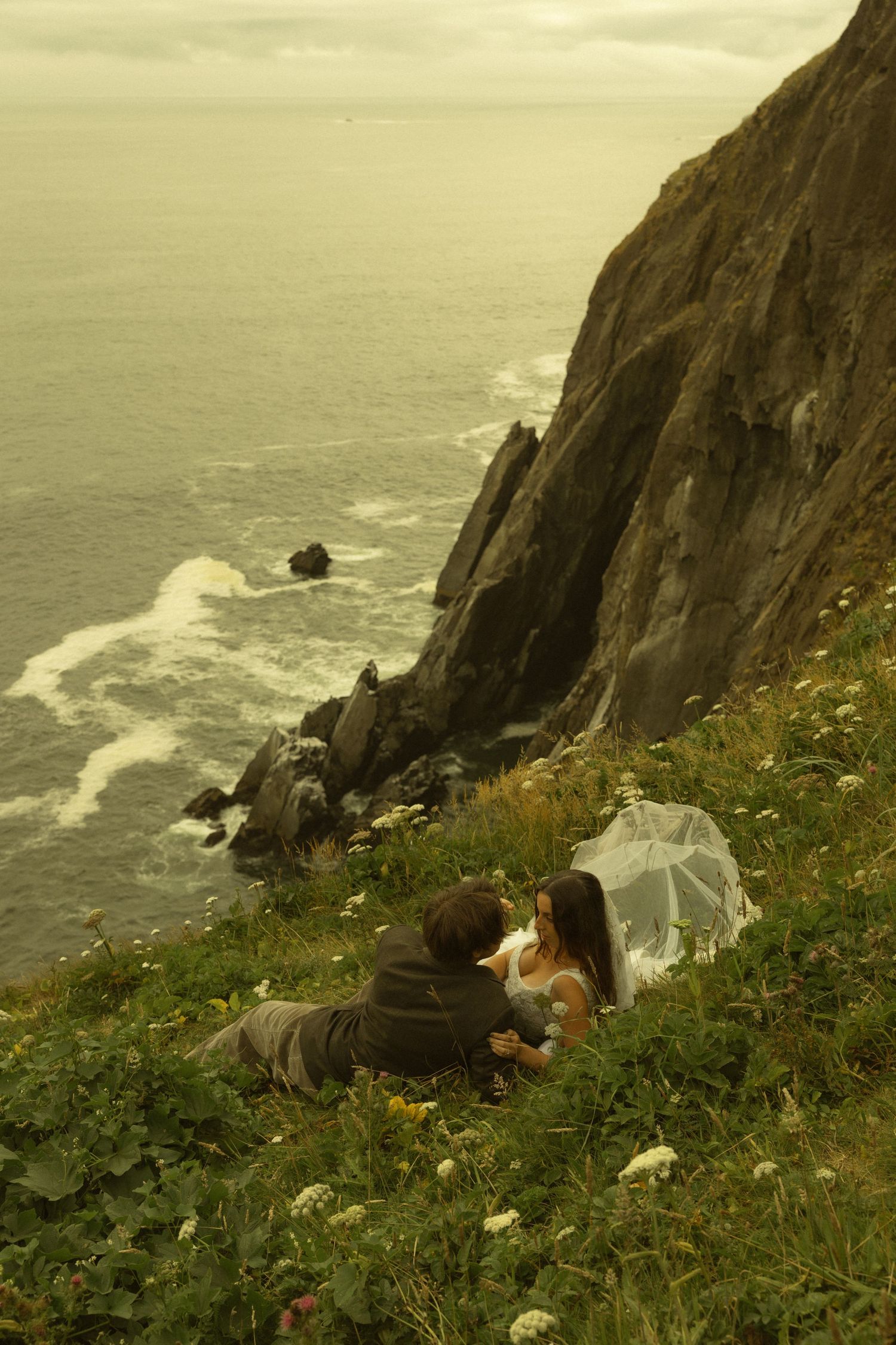 A couple in wedding attire sits on a cliffside meadow overlooking a dramatic rocky coastline and ocean.