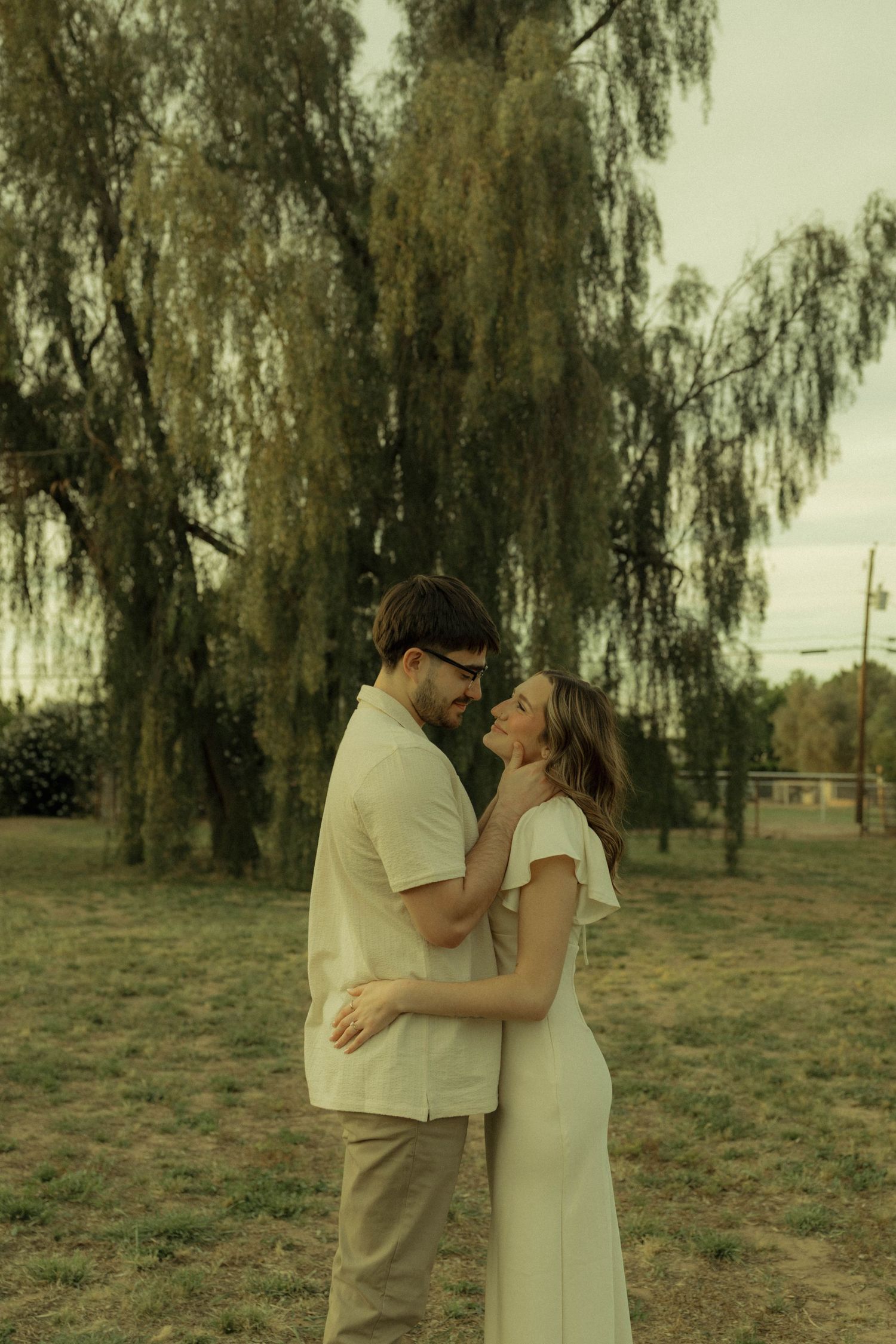 Couple smiling and holding each other tenderly under a weeping willow tree.