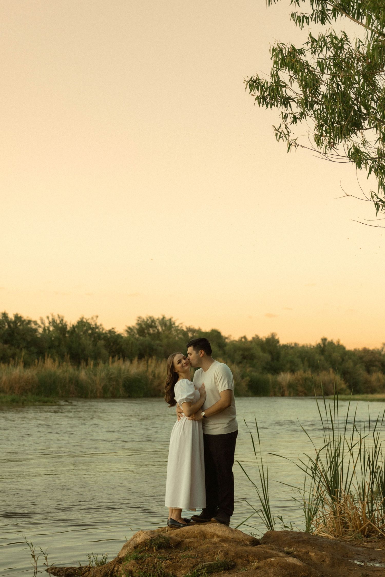 Loving couple hugging closely on a rock by a calm river at golden hour with trees and reeds nearby.