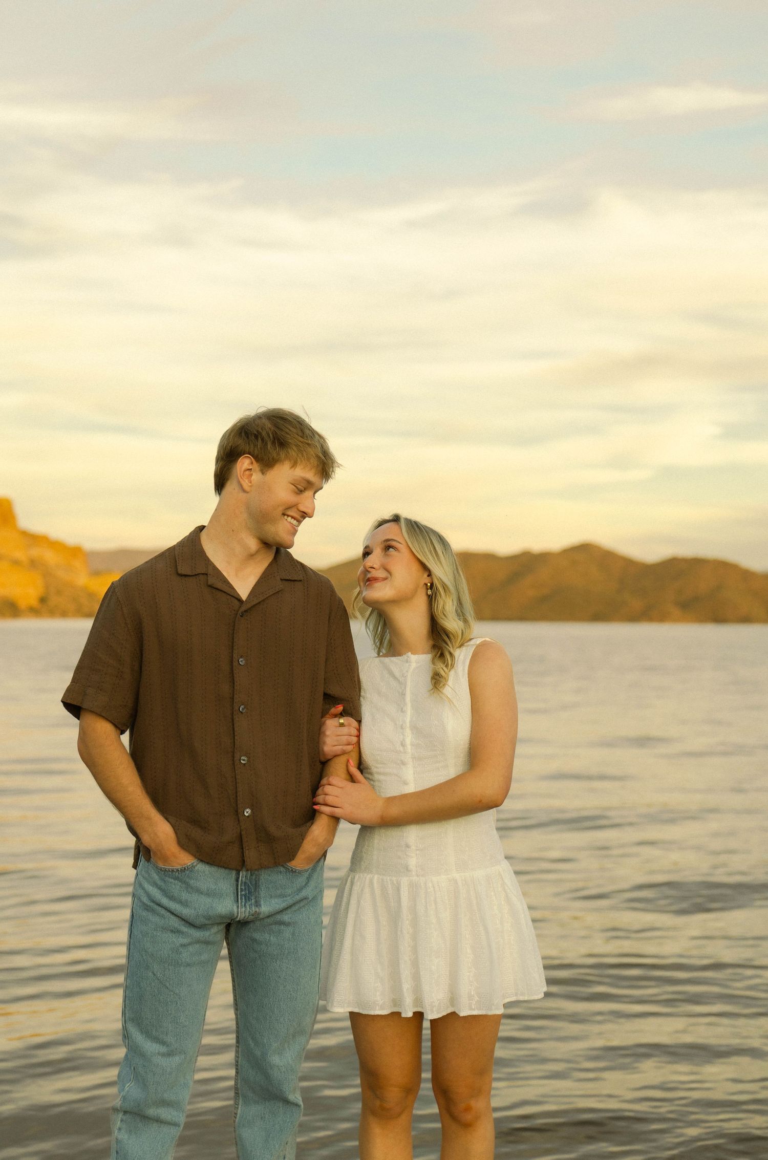 A romantic couple shares a tender moment at sunset by a lake with mountains in the background.