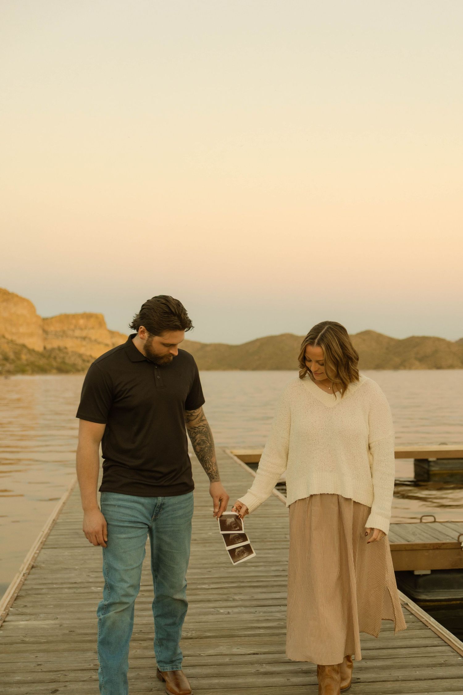 A couple walks on a wooden dock holding ultrasound photos during golden hour by a lake.