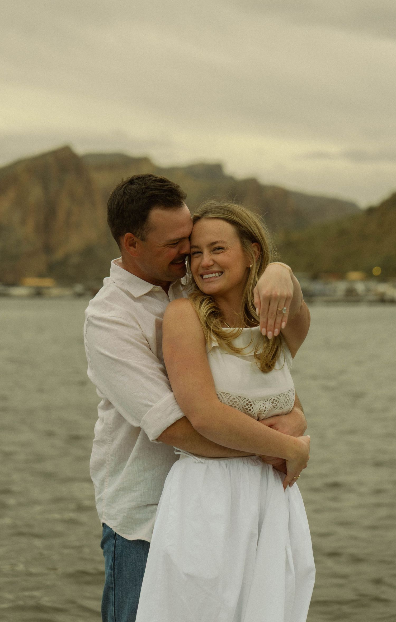 A joyful couple shares a loving moment while embracing by the water with mountains in the background.