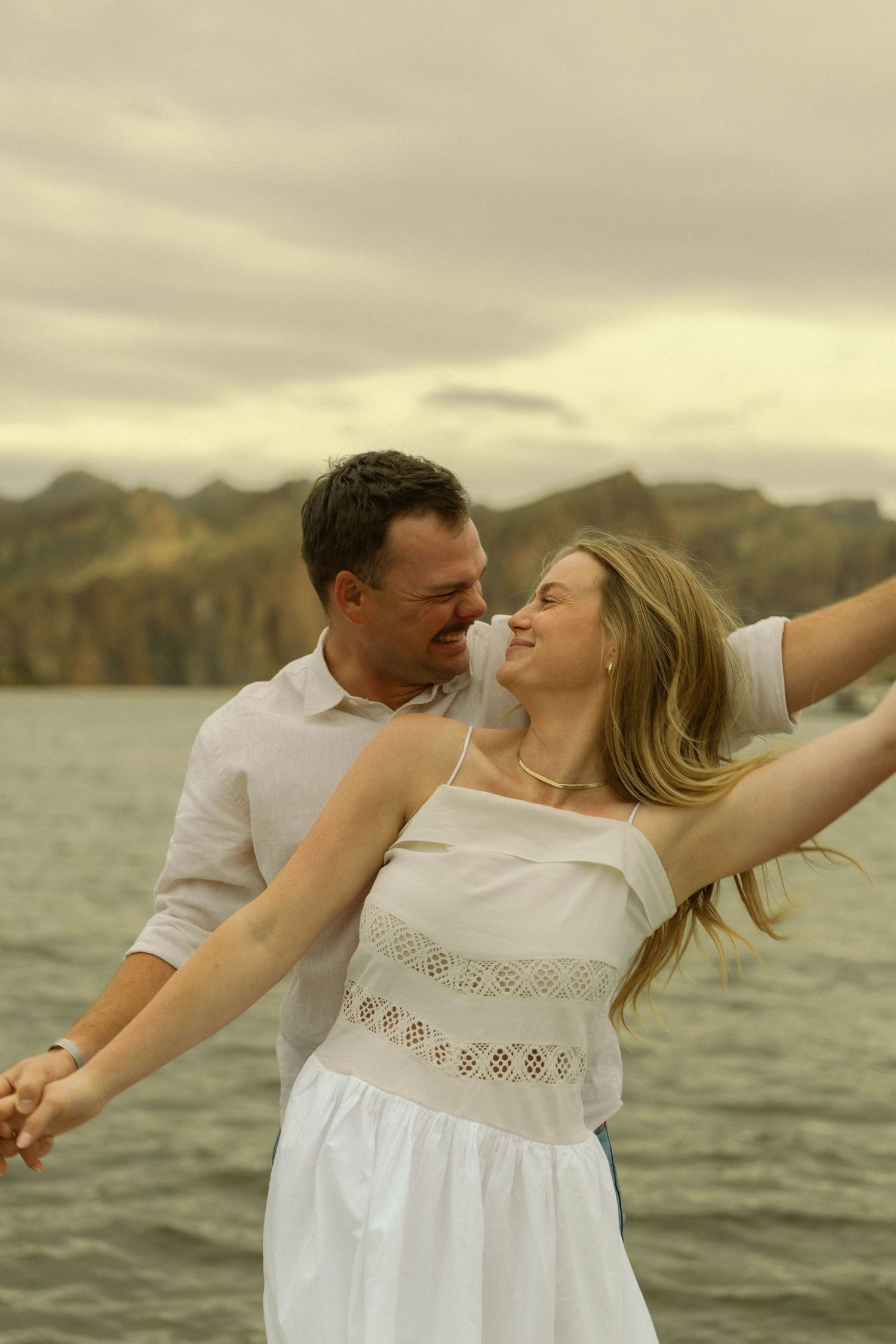 A romantic moment captured at sunset by the water with a couple in white clothing embracing against a mountain backdrop.