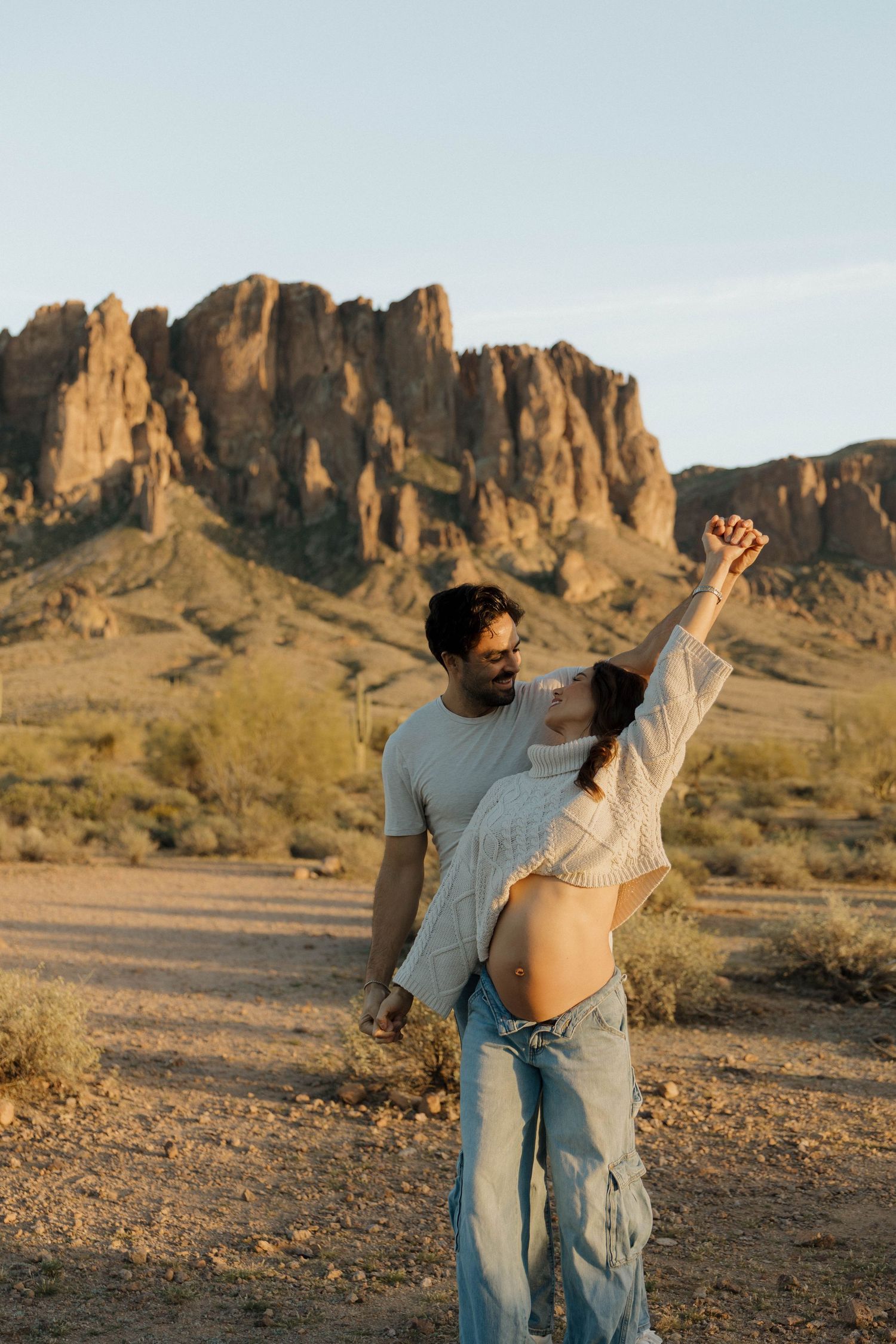 A couple dances playfully against the backdrop of rugged desert mountains during sunset.