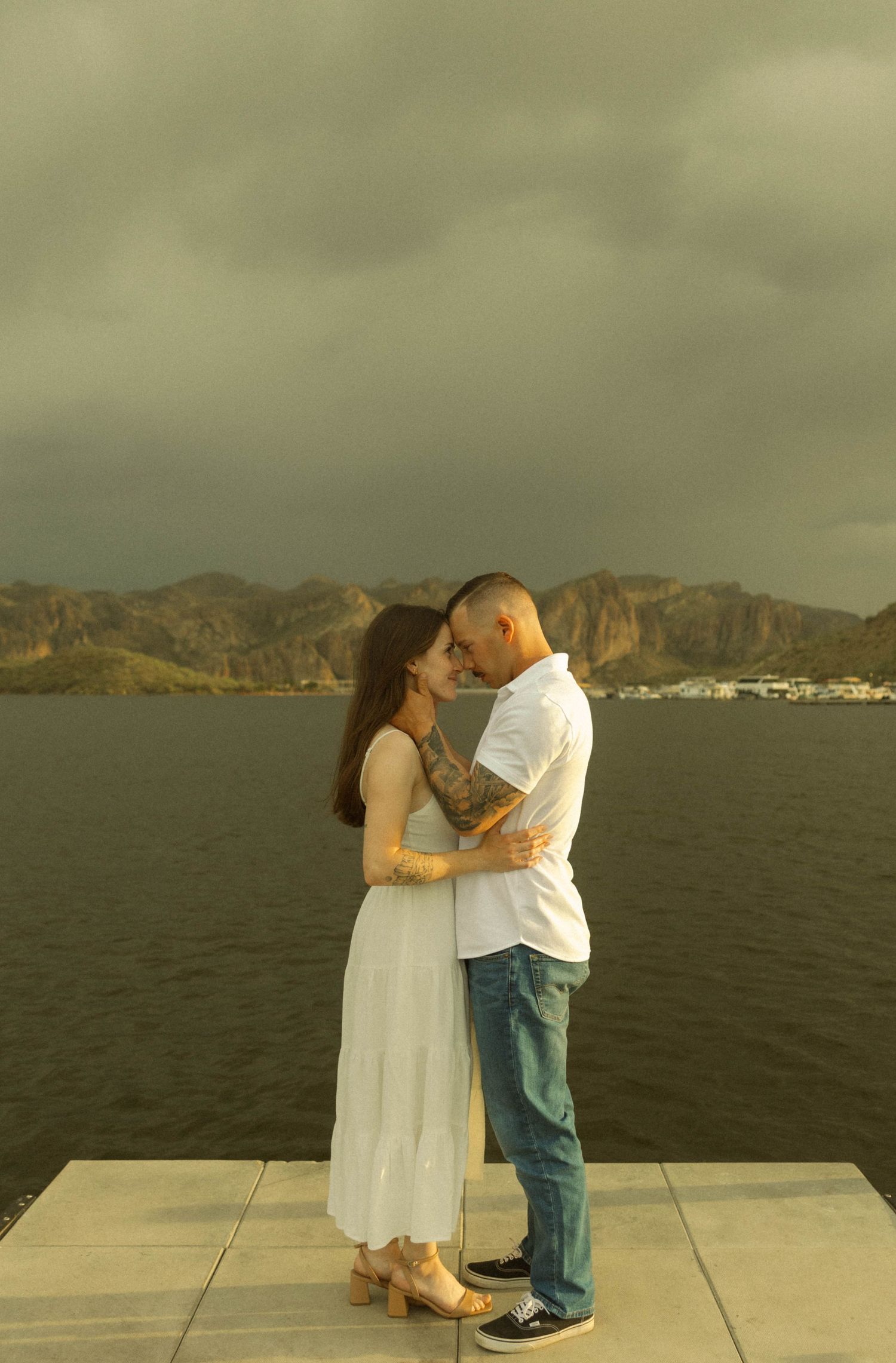 A couple shares romantic moments on a seaside platform with dramatic mountains and ocean views in the background.