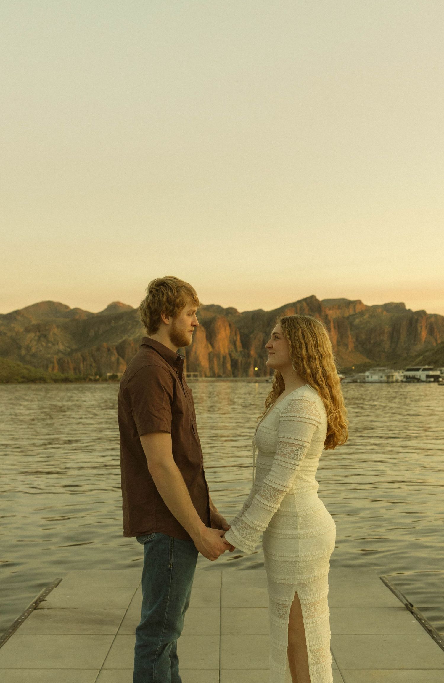 Couple holding hands on dock by lake with mountainous landscape at sunset.