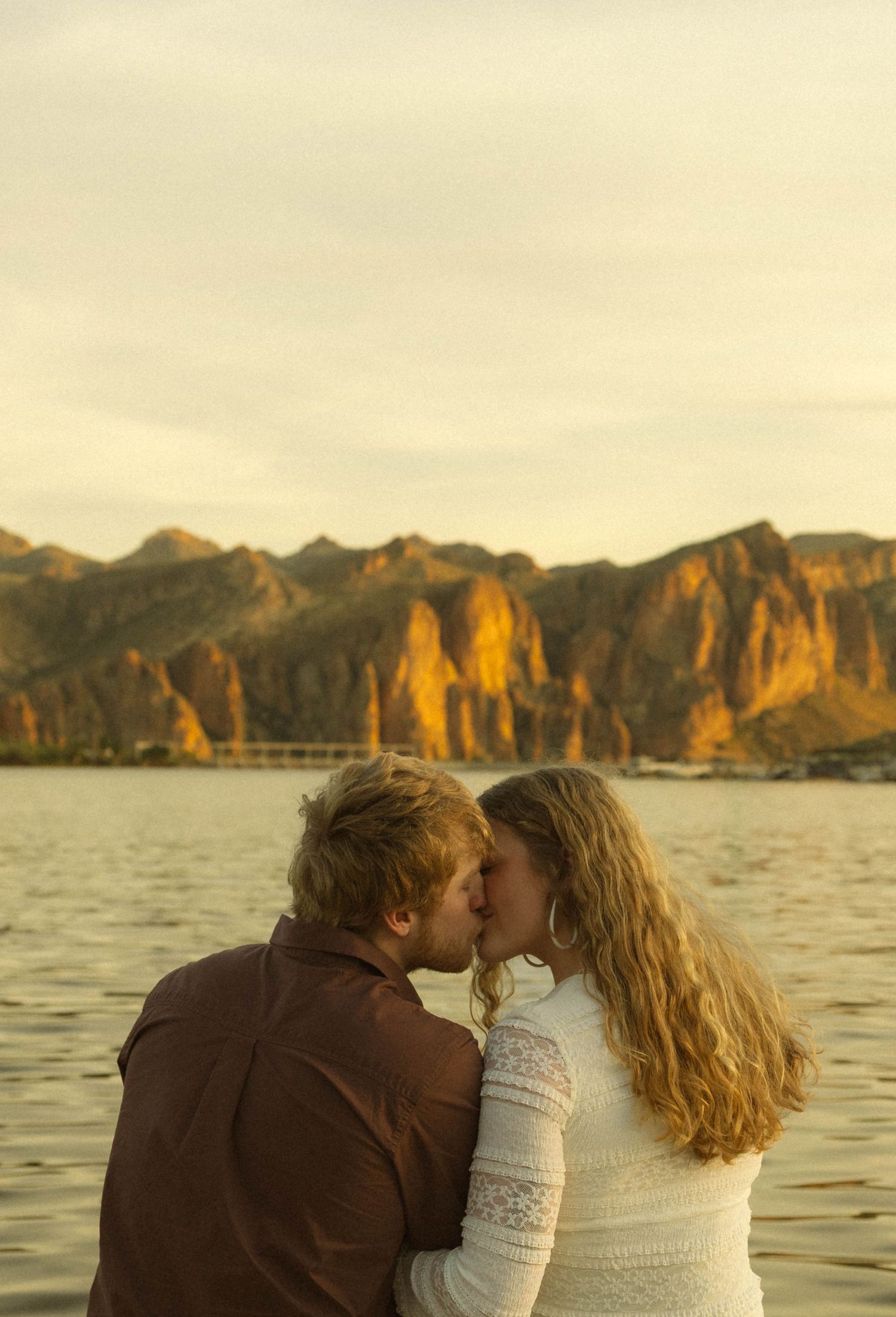 A romantic couple shares a tender moment at sunset by a lake with dramatic mountain cliffs in the background.
