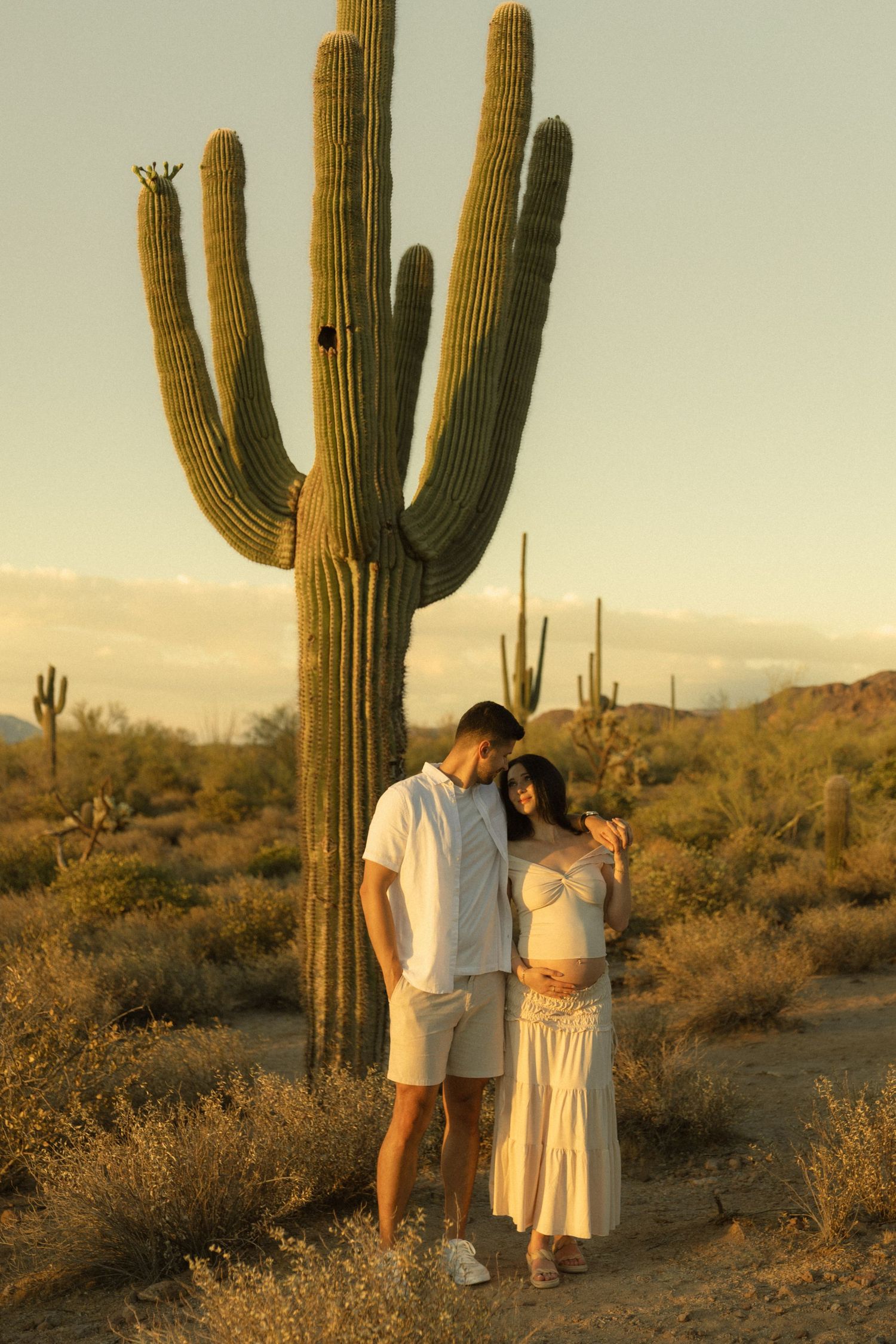 Couple in white outfits posing before a tall saguaro cactus in the Arizona desert at golden hour.