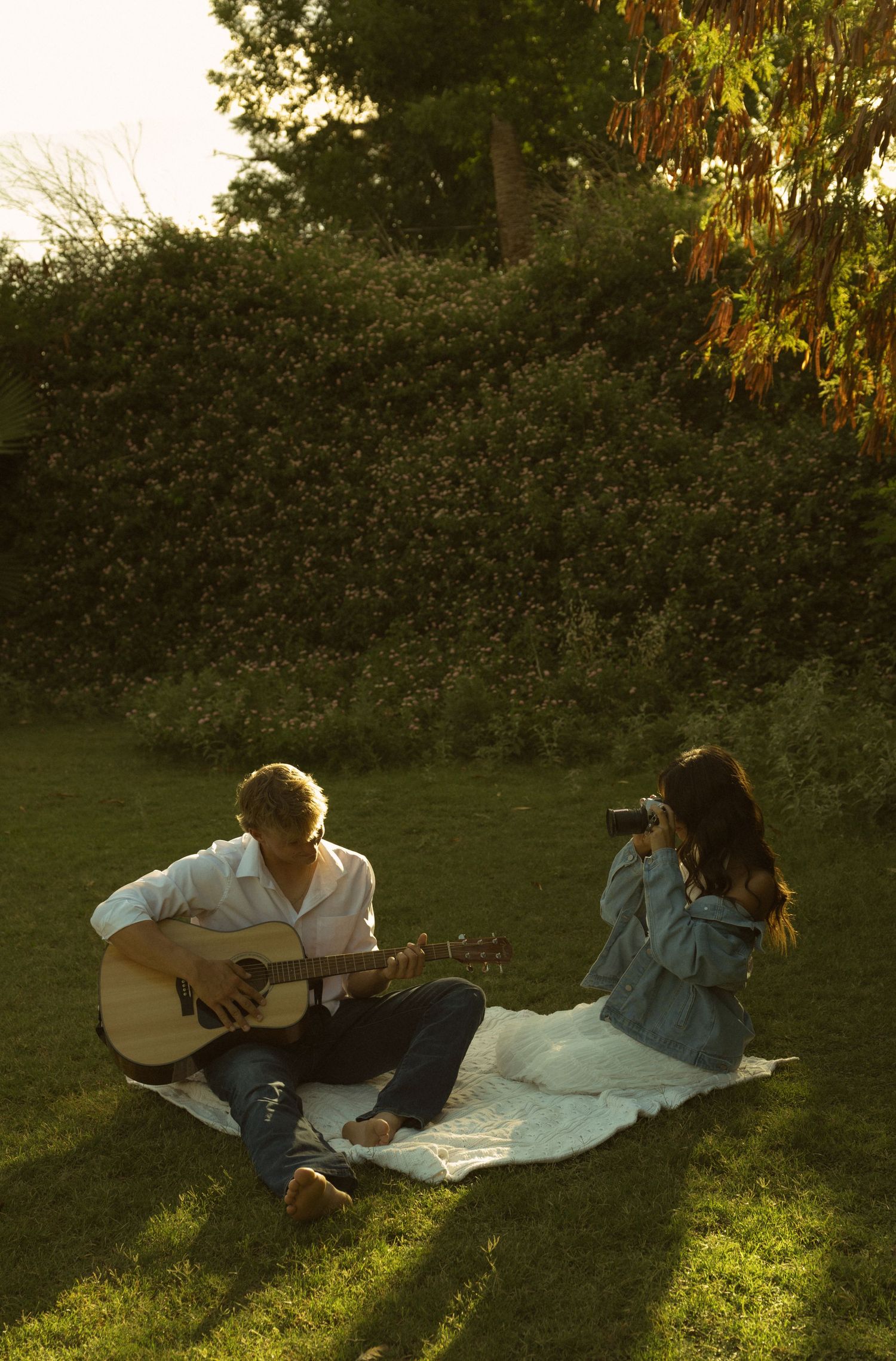 Couple sitting on blanket in garden at sunset, man playing acoustic guitar while woman photographs him.