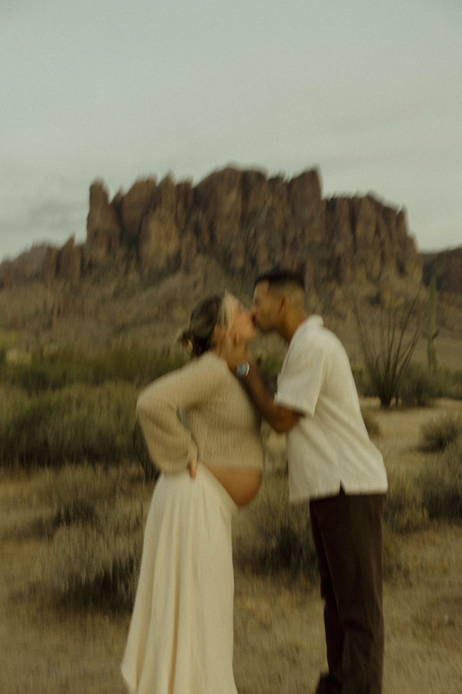 A couple shares a romantic kiss in front of Superstition Mountains in the Arizona desert during a maternity photo session.