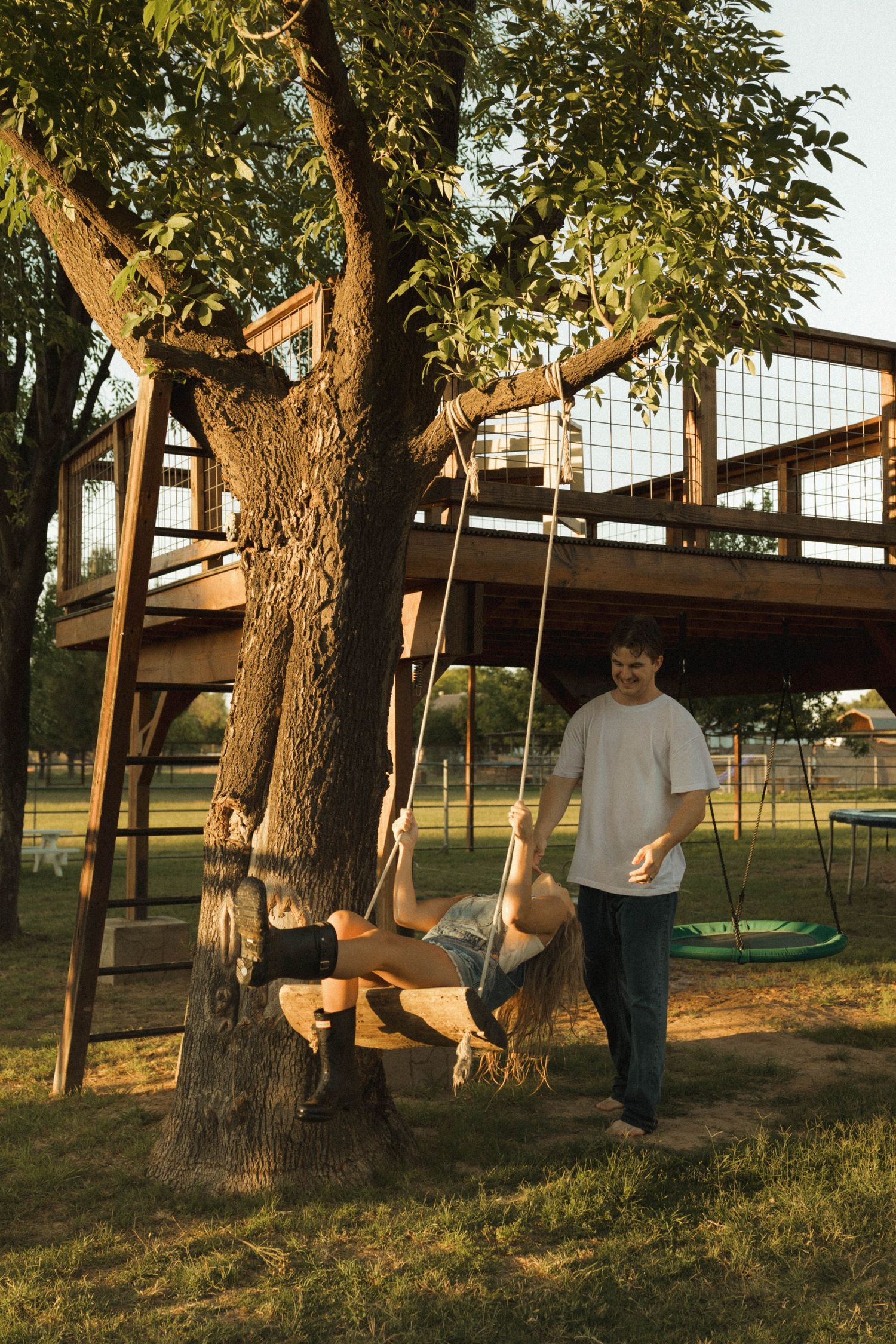 A man pushes a woman on a wooden tree swing beneath a treehouse in a sunlit backyard.