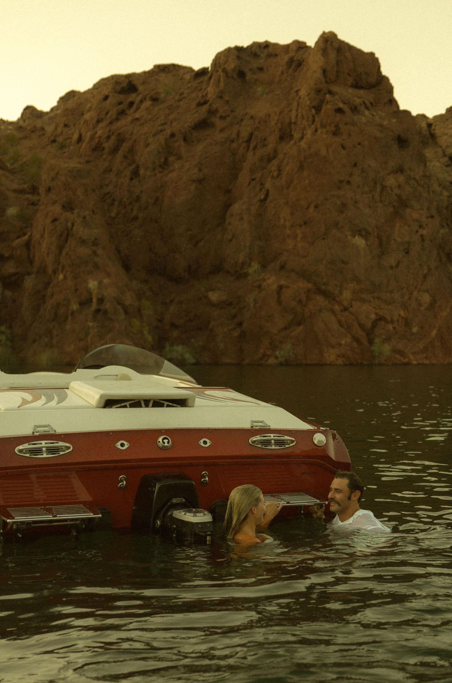 Couple swimming beside a red vintage speedboat on a calm lake with dramatic red rock cliffs in the background.
