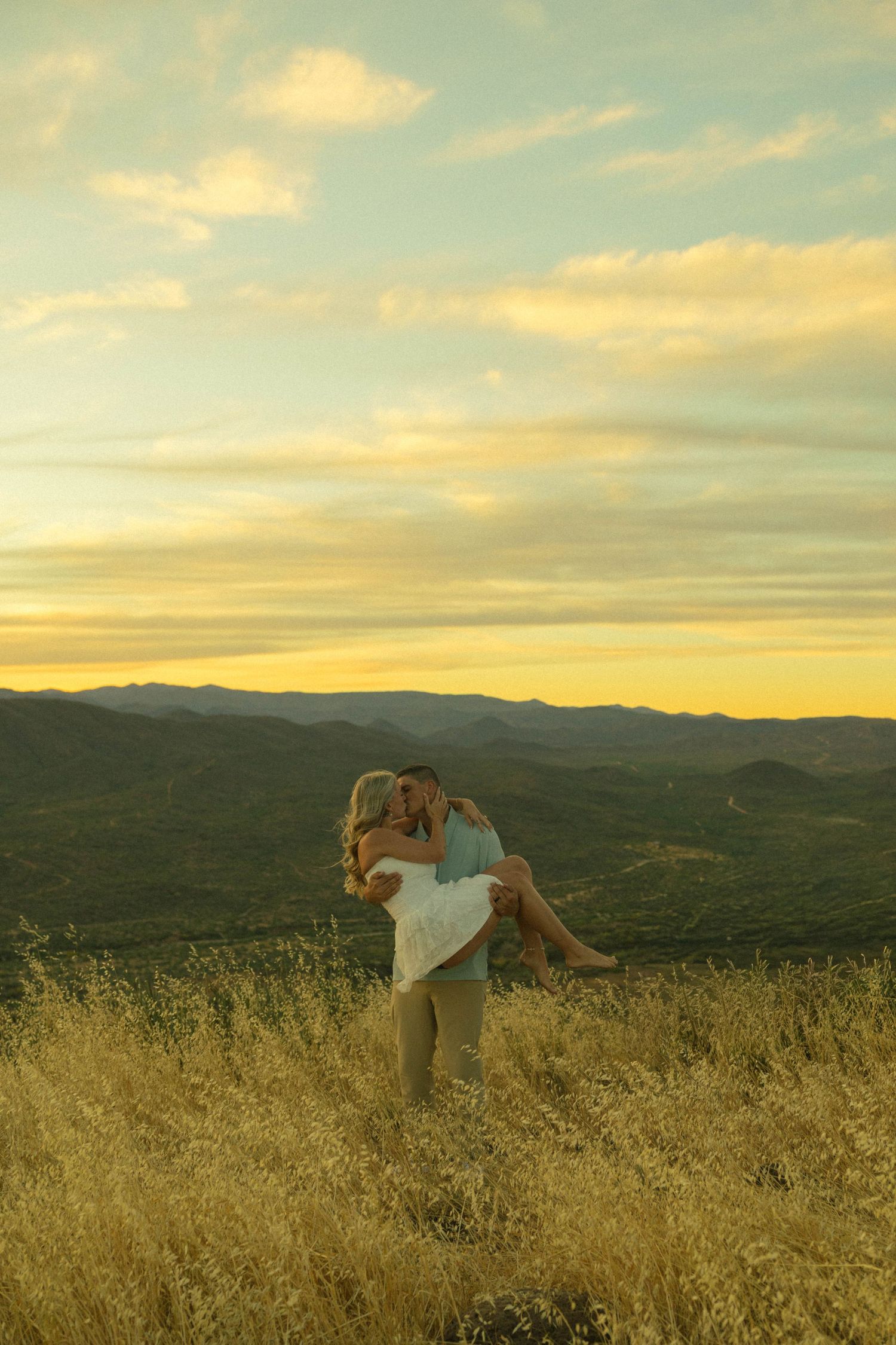 Man lifting woman in white dress for a kiss at sunset over a golden mountain landscape.