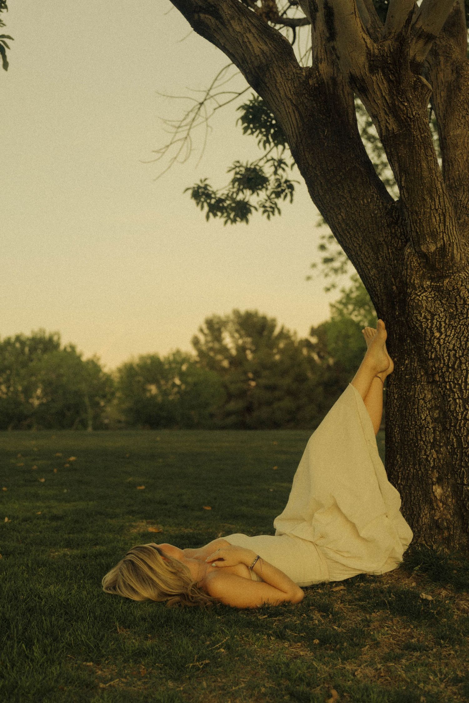 Woman in white dress lying on grass with legs resting against a tree at sunset.