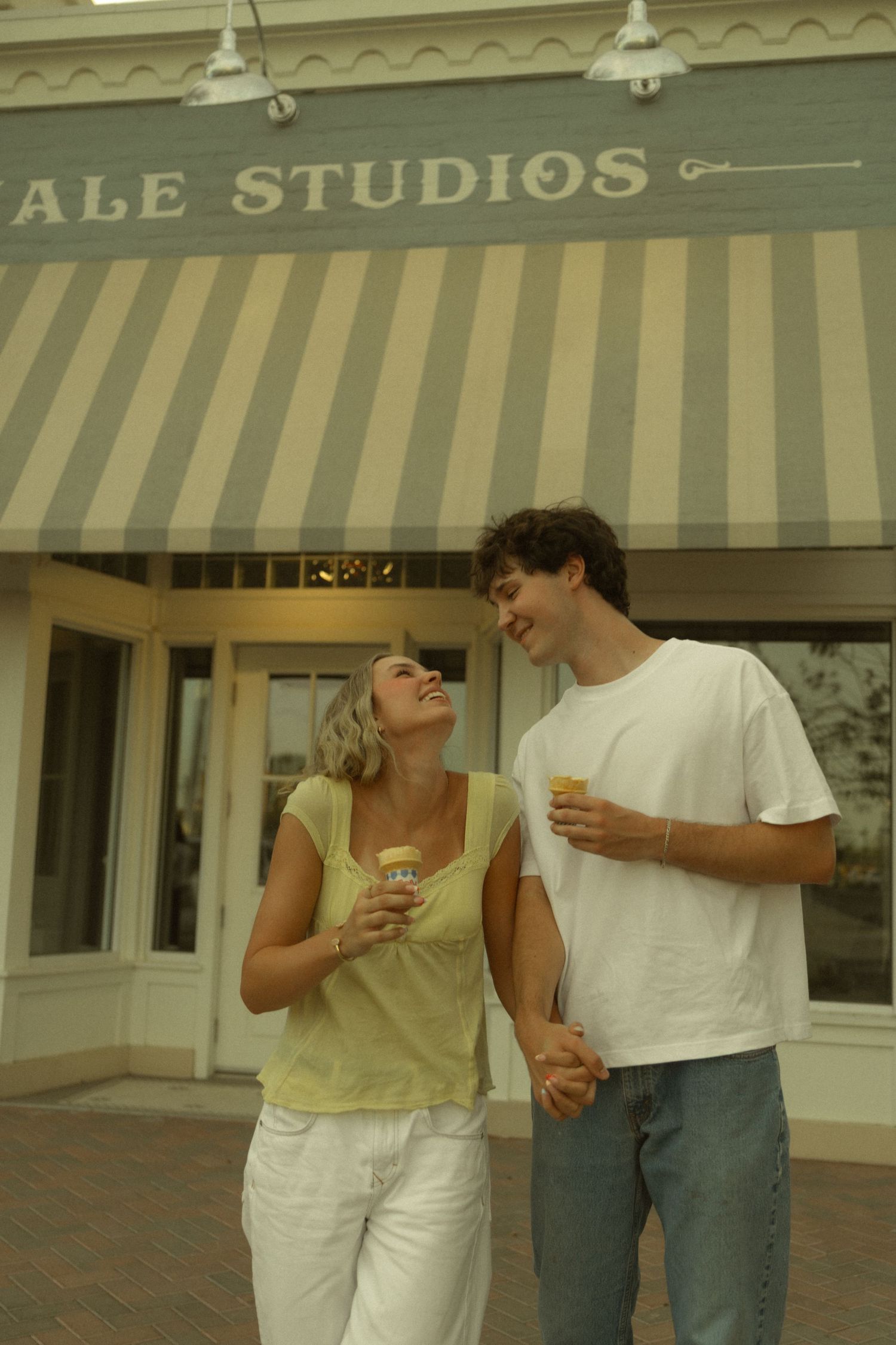 Young couple laughing and holding ice cream cups outside a studio storefront with striped awning.