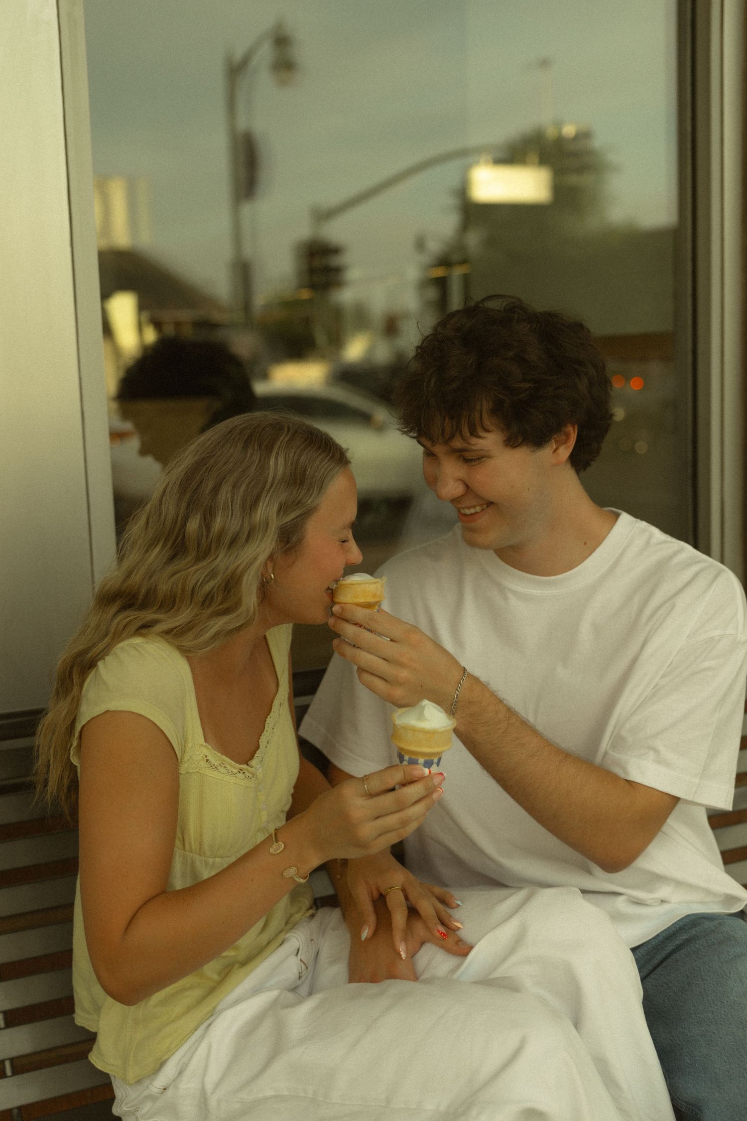 Young couple sharing ice cream cones on a bench outside a shop, smiling and enjoying a sweet summer date.