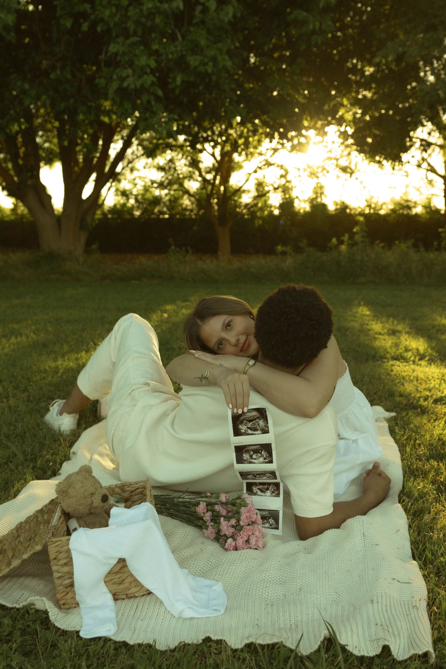 Couple sitting on blanket in park at sunset, holding ultrasound photos, sharing tender embrace among pink roses and teddy bear.