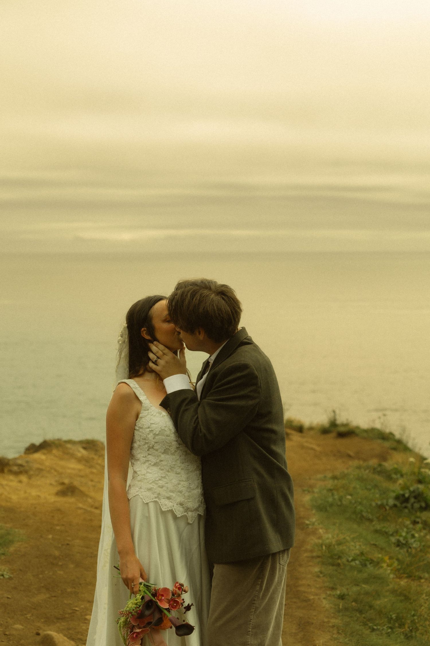 Bride and groom share a romantic kiss on a coastal cliff at sunset, bride holding a colorful bouquet in a white gown.