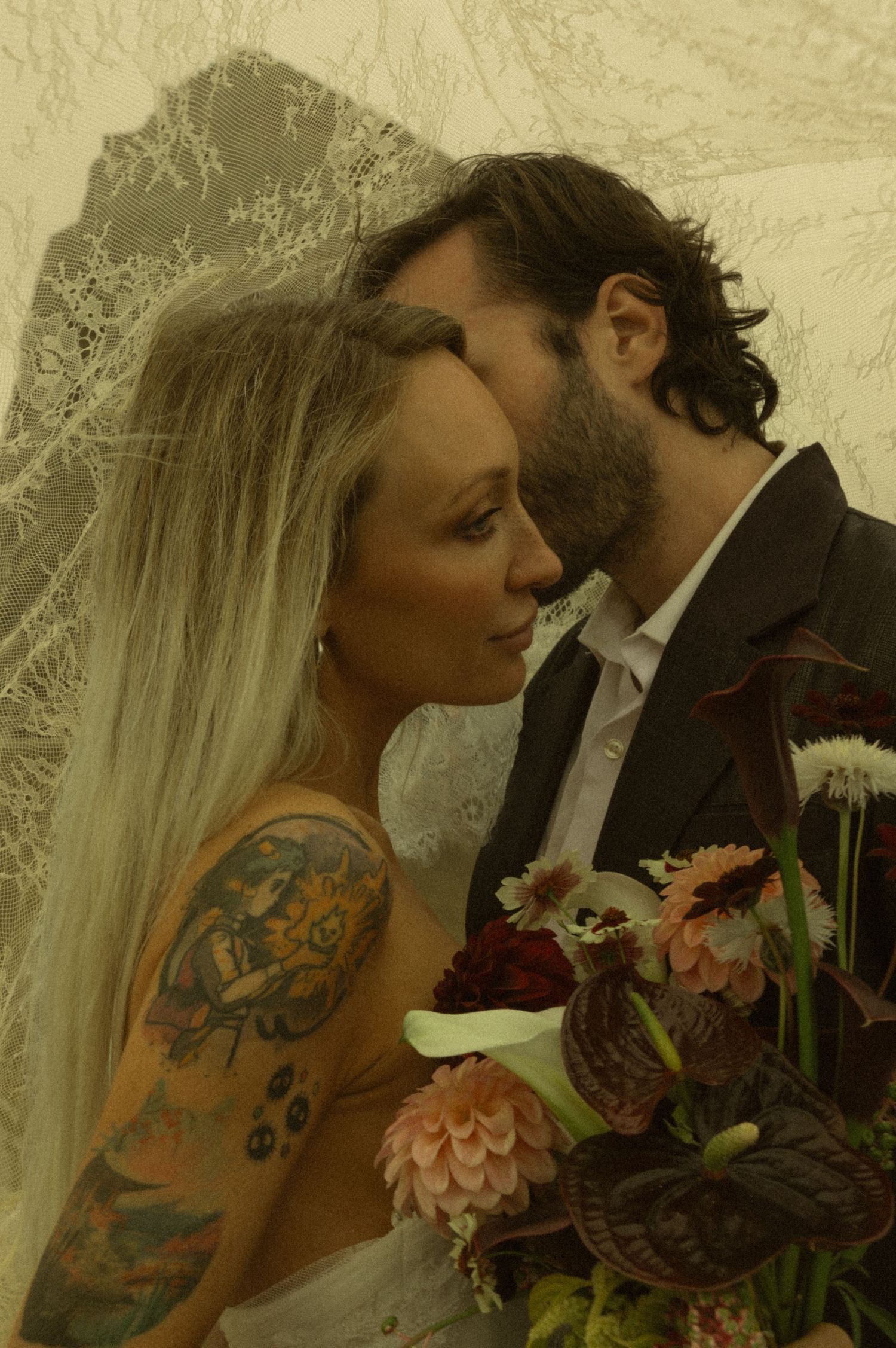 Tattooed bride and groom sharing an intimate moment under lace veil, holding dark floral bouquet with dahlias and anthuriums.