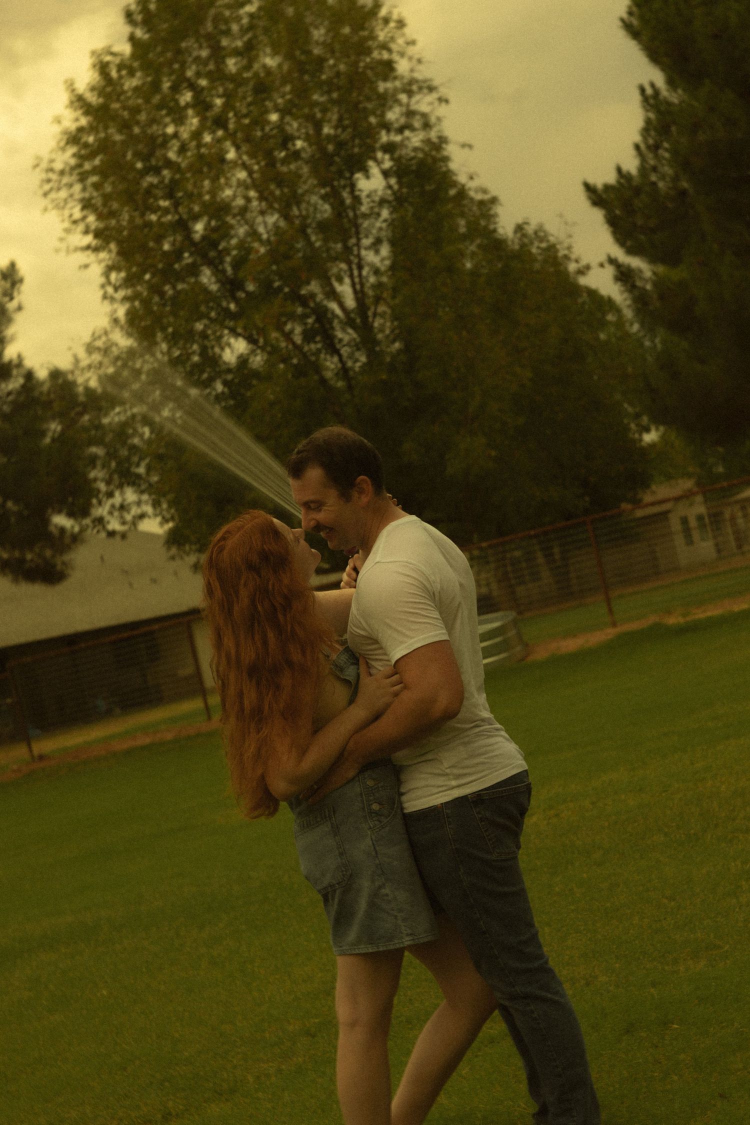 Couple embracing and dancing on green grass in a backyard with trees and a sprinkler in the background.