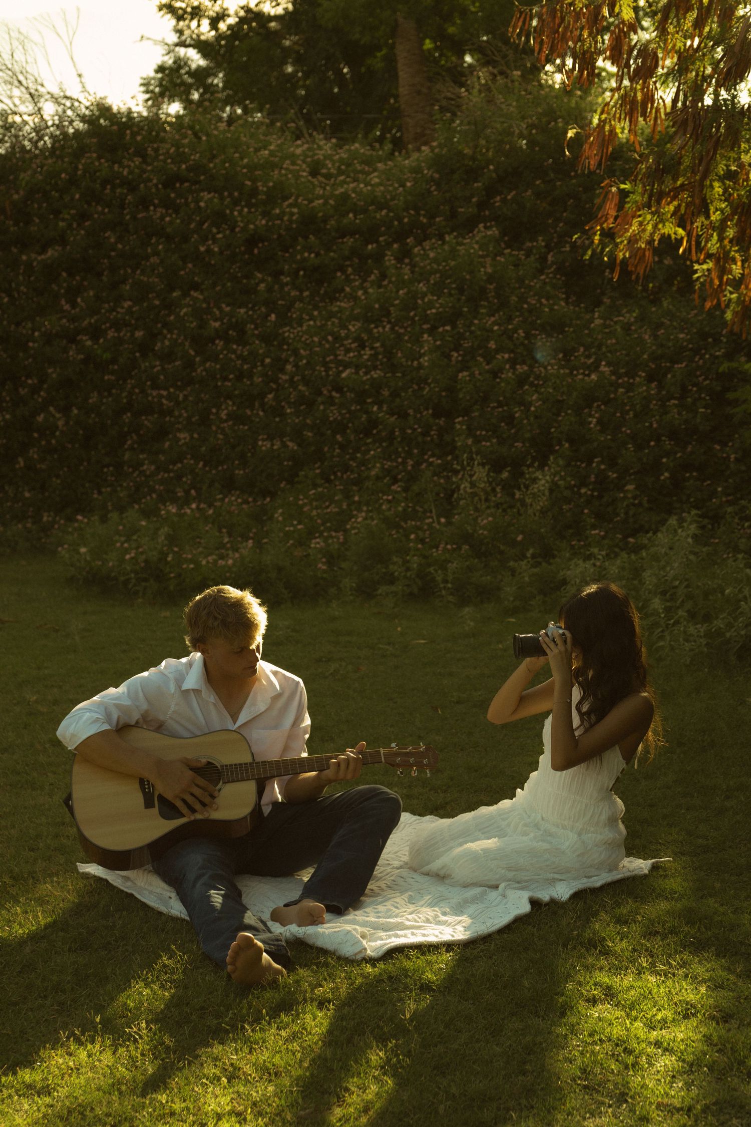 Couple enjoying a golden hour picnic on grass, man playing acoustic guitar while woman photographs him with a vintage camera.
