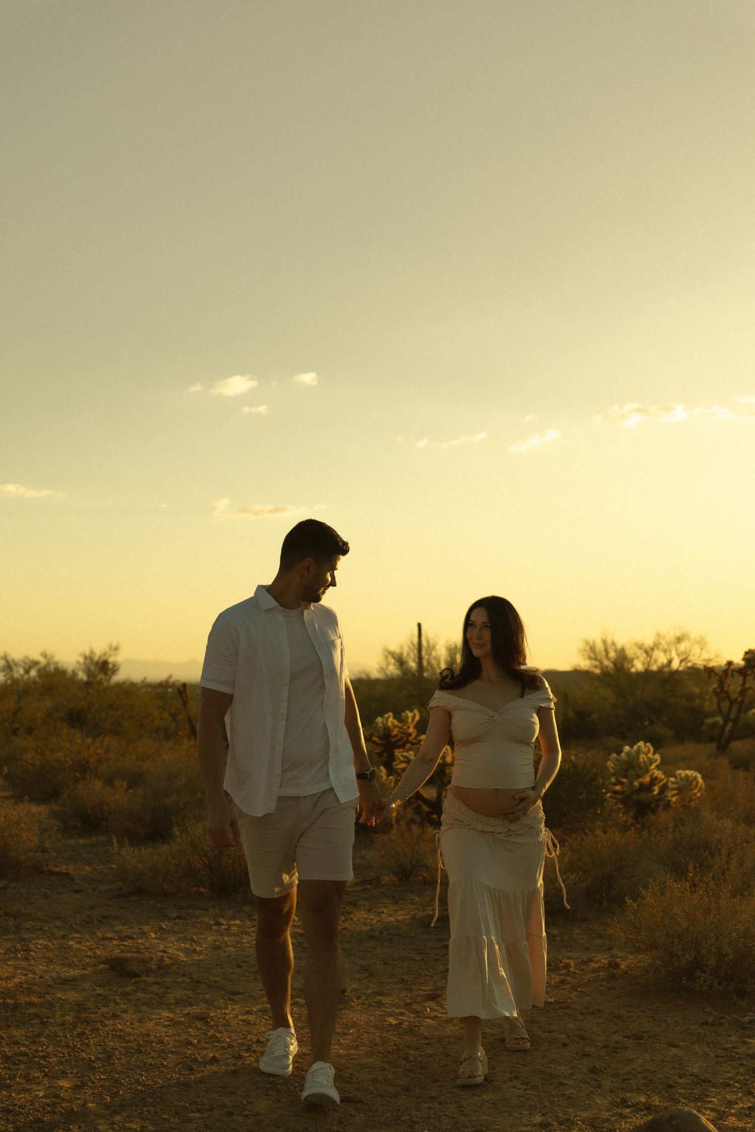Pregnant couple holding hands at golden hour in desert landscape, woman in white dress cradling baby bump.