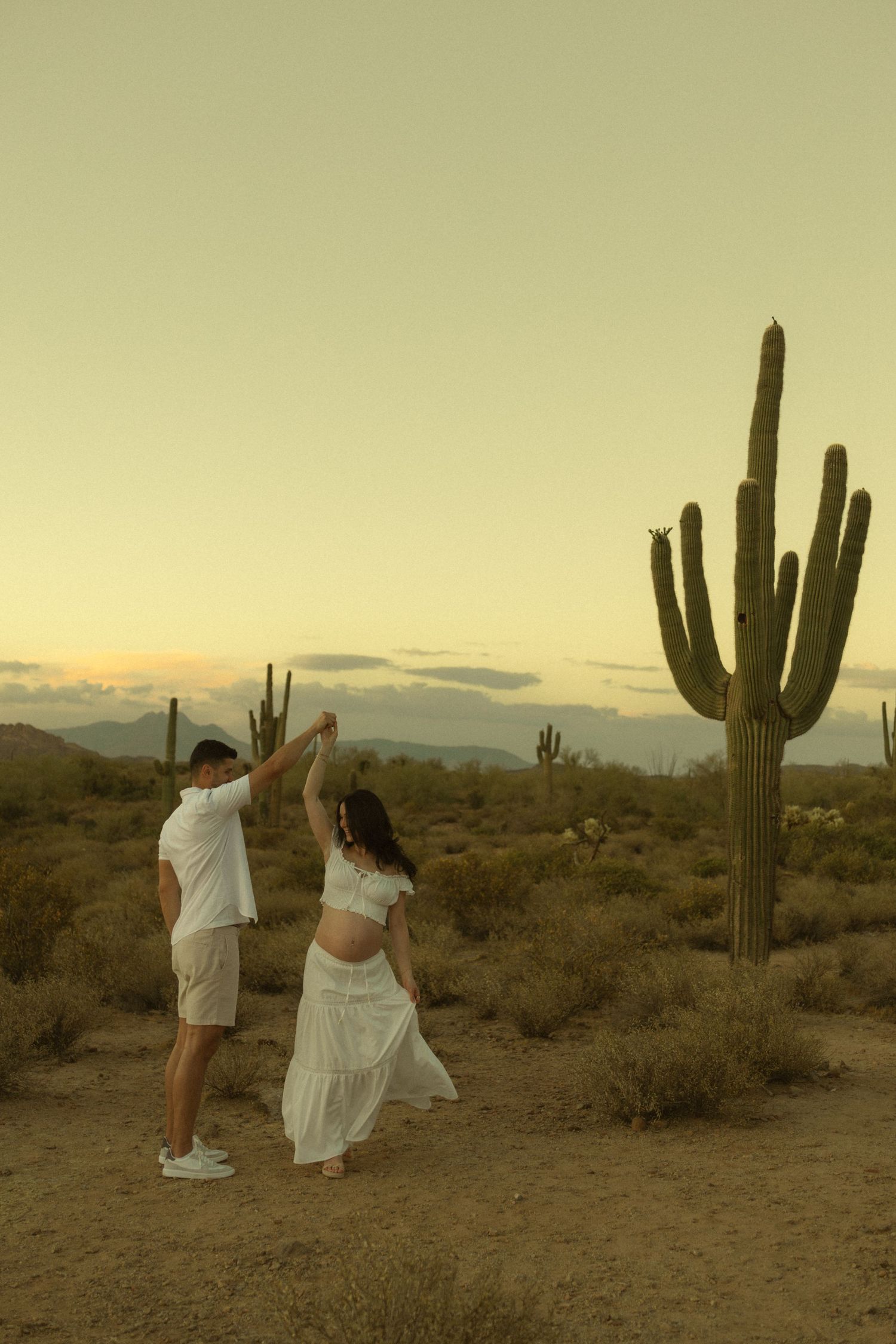 A couple dances together in the Sonoran Desert at sunset, with towering saguaro cacti and mountains in the background.